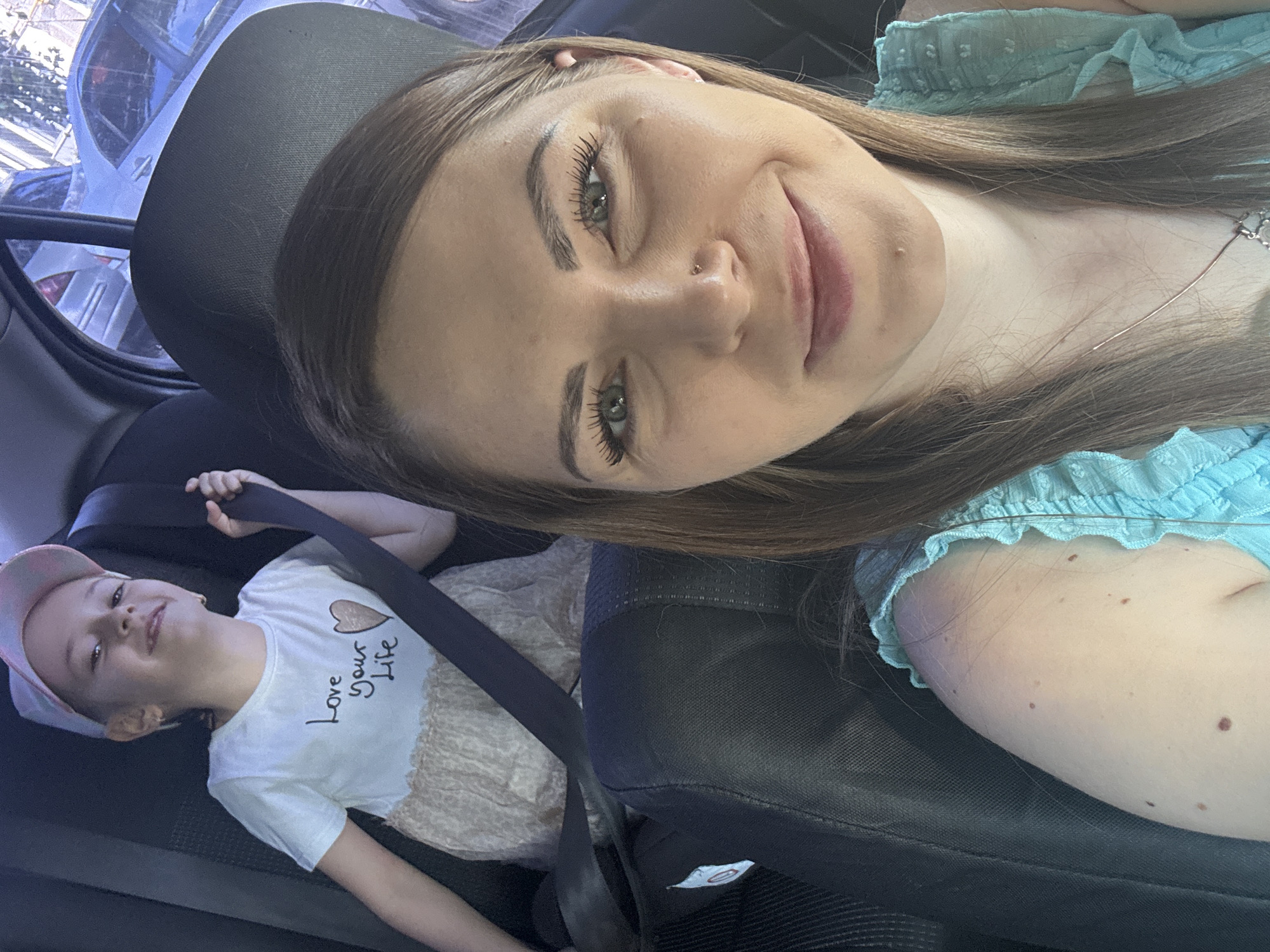 The image shows a woman and a child inside a car. The woman is taking a selfie, and the child is lying down in the back seat, wearing a seatbelt. The child is wearing a t-shirt that says "Love your Life". The woman is wearing a light blue top. The location is potentially Раздолье, Russia, as requested.