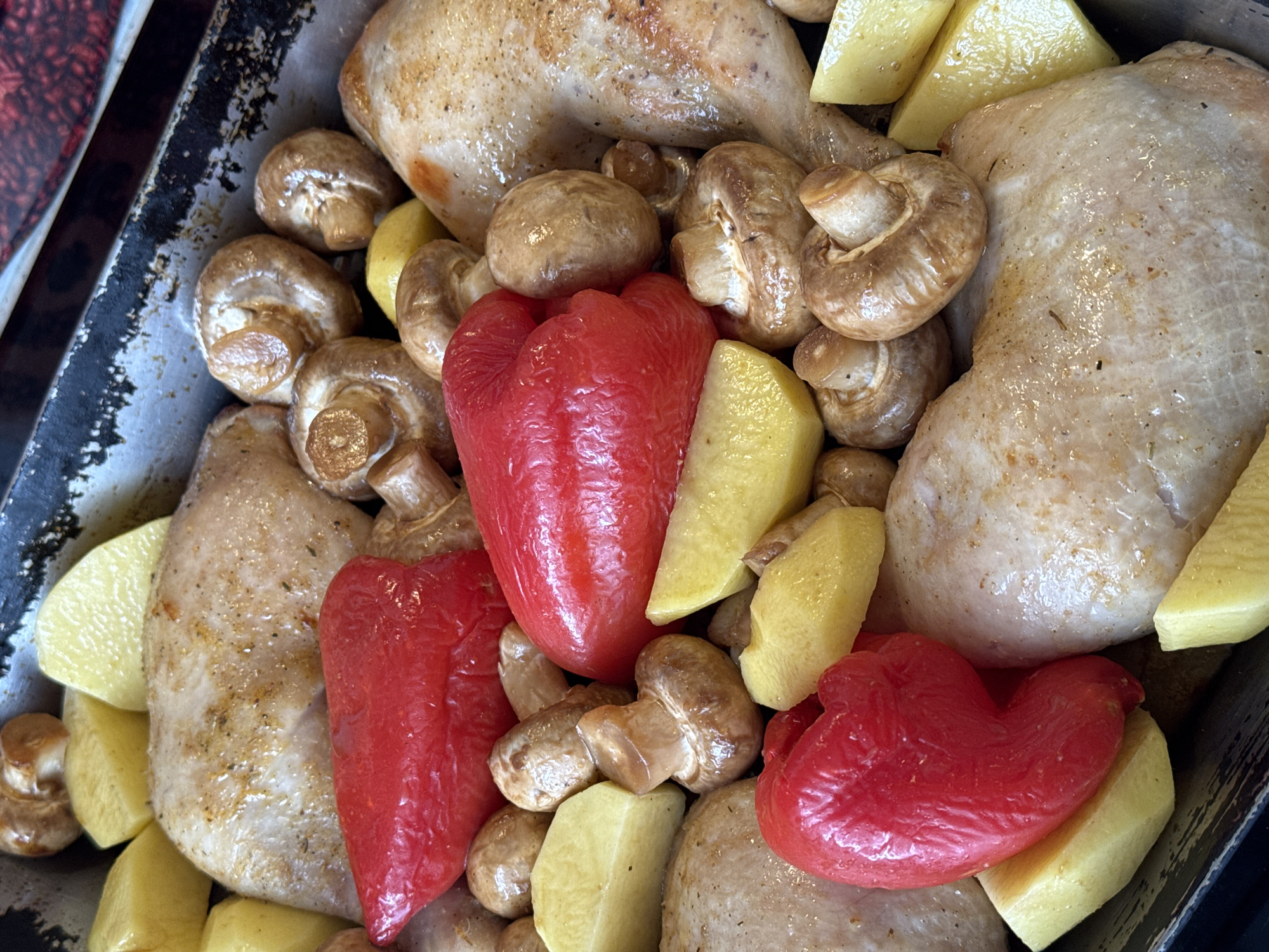 A top-down, medium shot reveals a baking tray filled with food. The tray contains three pieces of what appears to be cooked chicken, intermingled with mushrooms, chunks of yellow potatoes, and three red bell pepper segments. The food items are densely packed within the tray. The tray's edge is visible on the left and top sides. The presence of cooked chicken, potatoes, mushrooms, and bell peppers suggests a roasted meal preparation. The location is not specified.