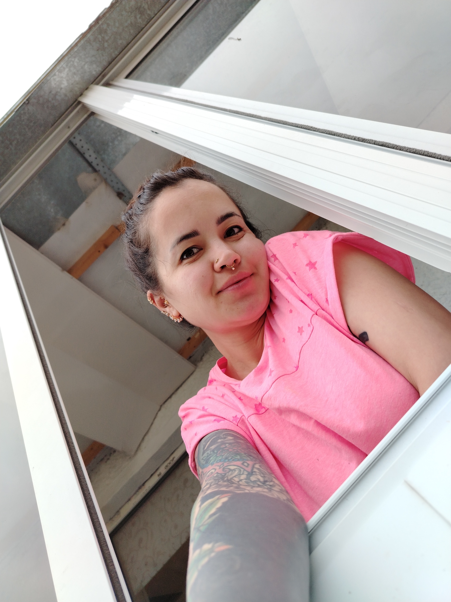 A female individual is depicted looking upward, positioned within a partially open white PVC window frame. Her dark hair is pulled back. She has brown eyes, a slight smile, a gold-colored ring piercing in her left nostril, and multiple small gold-colored hoop and cuff piercings on her left ear. She is wearing a pink t-shirt with subtle star patterns. A dark, solid heart-shaped tattoo is visible on her right upper arm, and her left arm is adorned with a full sleeve tattoo featuring dark, intricate designs. A gray brush-like weather seal is present along the window frame. Beyond the window, the background reveals an exterior or semi-enclosed area, including a corrugated metal surface in the upper left, light-colored wall or ceiling sections, and wooden support beams and shadowed elements. This scene is located in Noviy, Russia.
