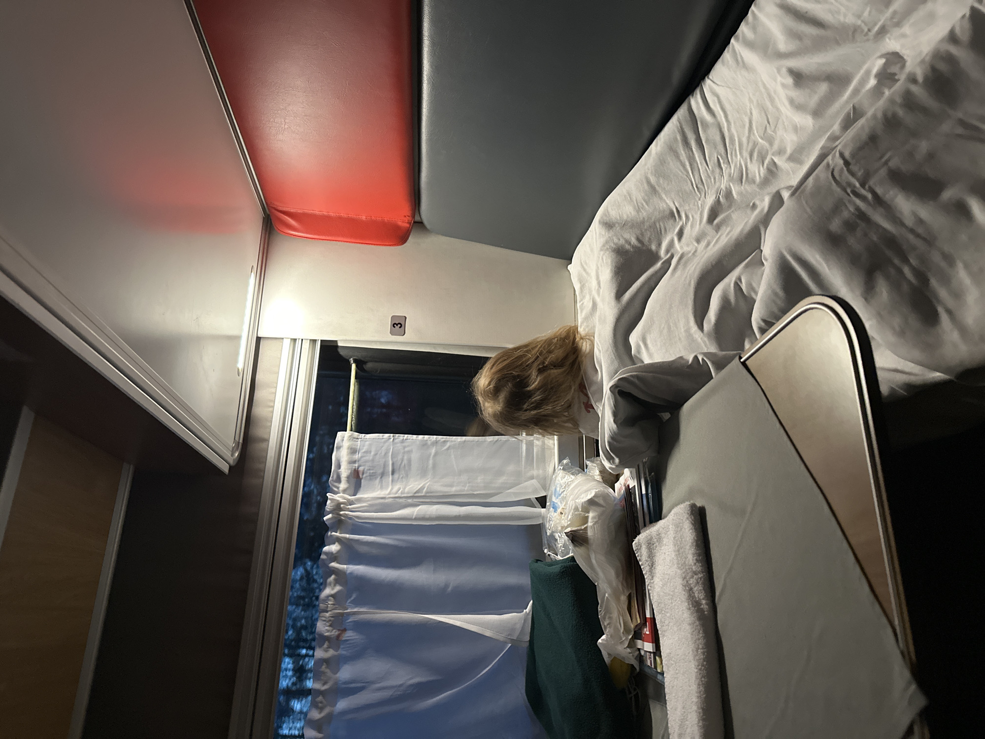 The image depicts the interior of a sleeping compartment, potentially in a train. A bed covered in white linens is visible on the right. Above the bed, there is a grey upholstered headrest. Adjacent to the headrest is a red upholstered bench seat. On the left, there is a wall panel with a light fixture. A person with blonde hair is visible near a window covered with white curtains. There is a small table in the foreground and a small bag with a dark green piece of clothing. The location is unclear. The mention of Malaya Ven'ya, Russia is unsupported.