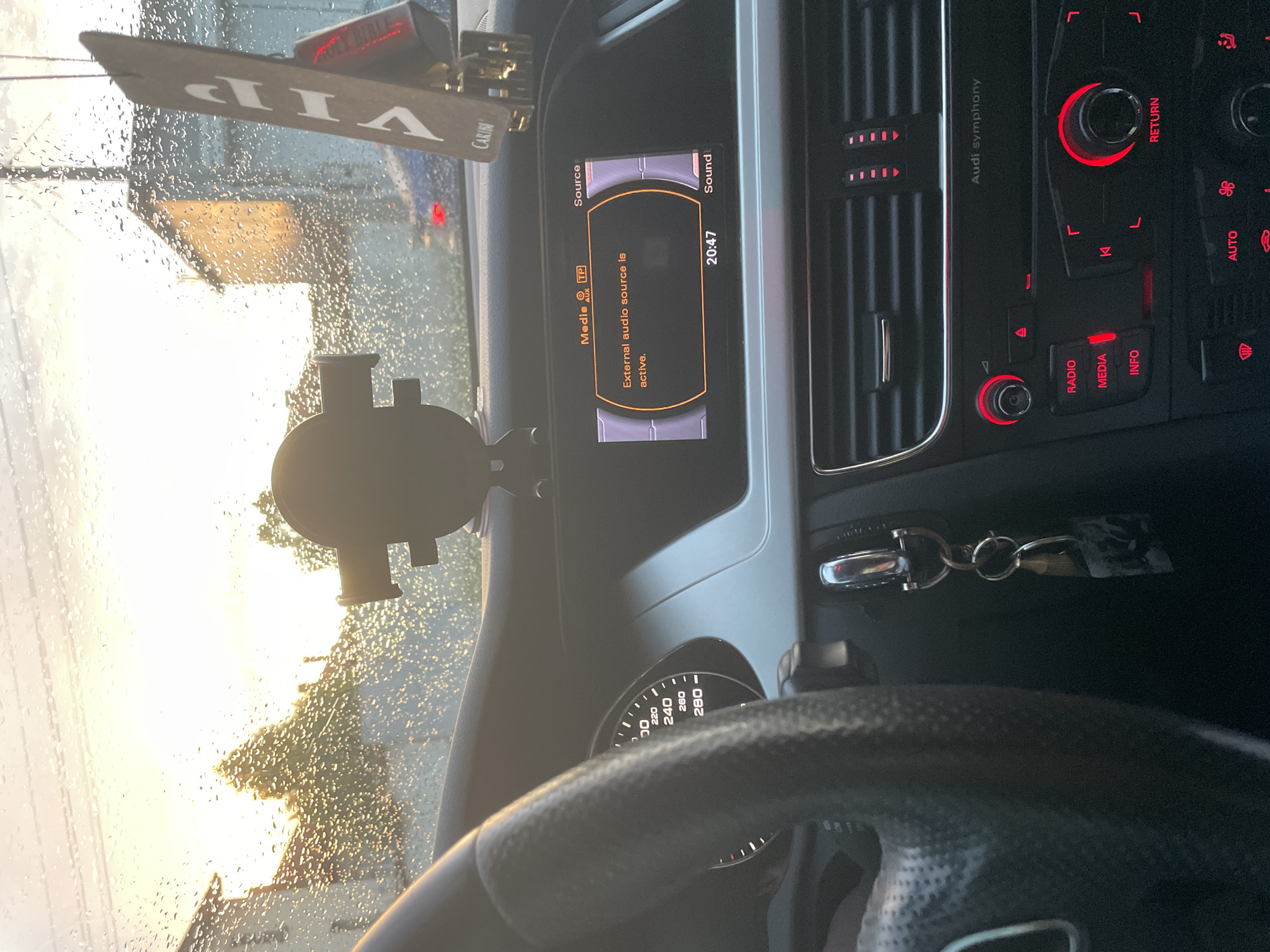 The image captures the interior of a car, specifically the dashboard and a view through the rain-splattered windshield. The scene is set inside an Audi, indicated by the "Audi symphony" text on the central console.
In the foreground, part of a black steering wheel is visible, with a speedometer showing readings up to 280 km/h partially seen through it. On the dashboard, a media display shows "Media AUX TP" at the top left, and a message in English: "External audio source is active." The current time, "20:47", is displayed on the screen, along with "Source" and "Sound".
To the right of the display, the car's control panel features red-illuminated buttons and dials for functions like "RADIO", "MEDIA", "INFO", "RETURN", and "AUTO", along with climate control vents. A keyring with keys hangs near the bottom right of the central display.
Hanging from the rearview mirror, an ornament or air freshener is visible, bearing the text "VIP" and "CARIBI". A smartphone holder is attached to the windshield in the lower left quadrant.
Outside the car, through the raindrop-covered windshield, a very bright, low-angle light, likely the sun setting or rising, illuminates the scene, casting a warm glow and creating silhouettes of distant trees or structures. The presence of numerous raindrops on the glass suggests recent or ongoing rain. The car is likely stationary, given the display and the angle of the shot. The geographical context provided is Ihtiman, Bulgaria.