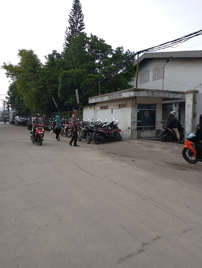 The scene shows a street in Katapang, Indonesia (latitude: -6.986, longitude: 107.555). A group of people are standing near a row of parked motorcycles. A man in a brown shirt is walking towards the camera, while a woman in a blue headscarf is standing next to a motorcycle. In the background, a white building with a grey roof is visible. Several power lines are strung across the street, and the building appears to be a business or industrial facility. The image was taken from a moving vehicle. The asphalt road is in good condition, with some minor cracks. There is green foliage and a tall tree on the left side of the image. The sky is a light grey. The sun is not visible. The image is taken at a slight angle.