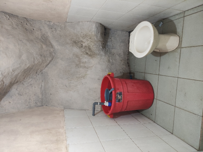 The scene is a small, tiled bathroom with a toilet and a red bucket. The walls are painted white and there is a small patch of concrete on the ceiling. The bucket is filled with water and contains a small blue container and a metal strainer. The toilet is white and sits on a tiled floor. The bathroom is in the Kecamatan Pujon area of Indonesia at coordinates -7.855 latitude and 112.454 longitude. There is no sign of people in the image.