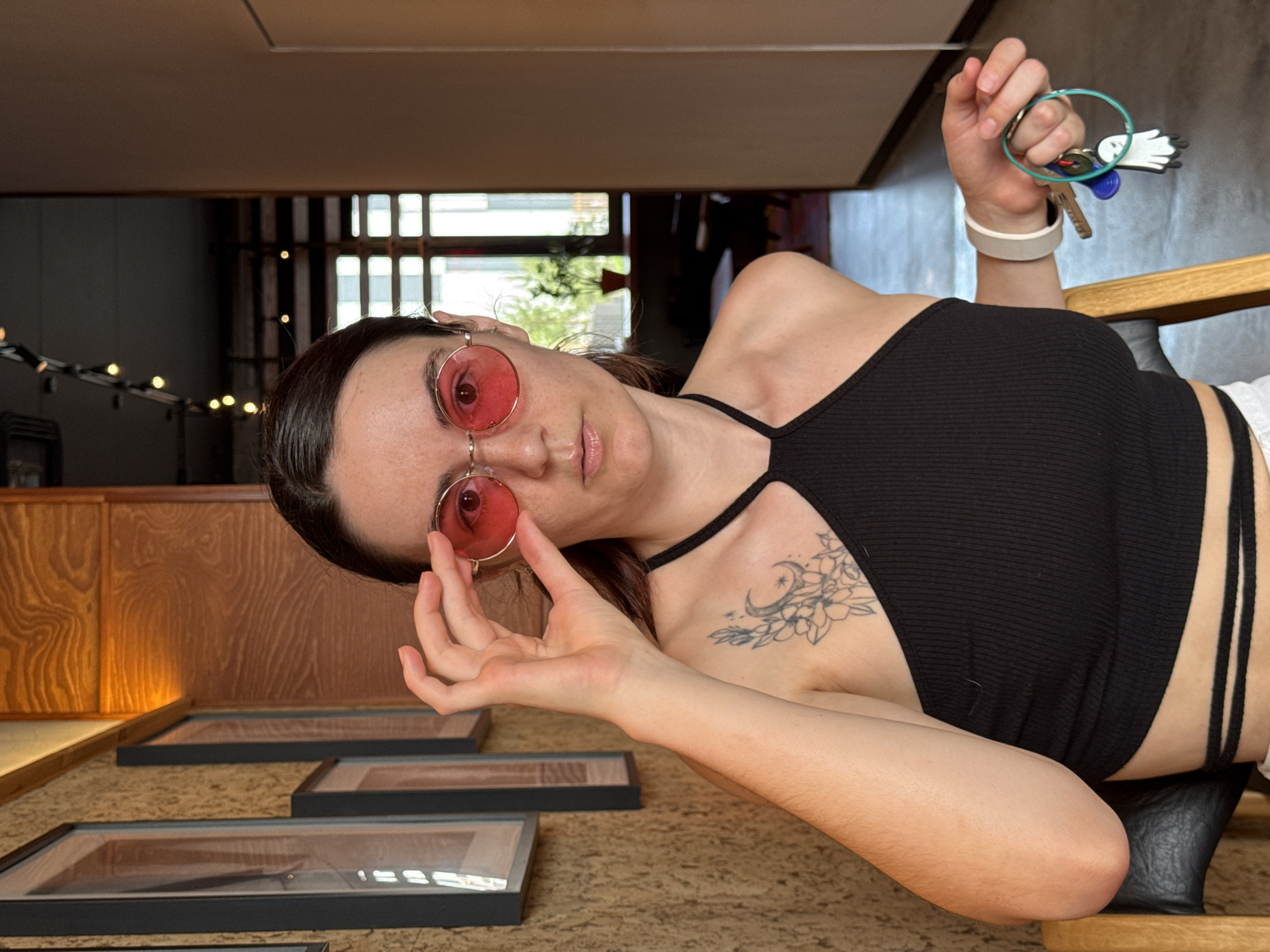 A woman with dark hair is looking at the camera. She is wearing pink sunglasses and a black halter top. She has a tattoo on her chest. She is holding keys in her right hand.
The background includes a wooden wall, framed objects, and window views. Based on the interior design and general location, this is likely a cafe or restaurant in Tyumen, Russia.