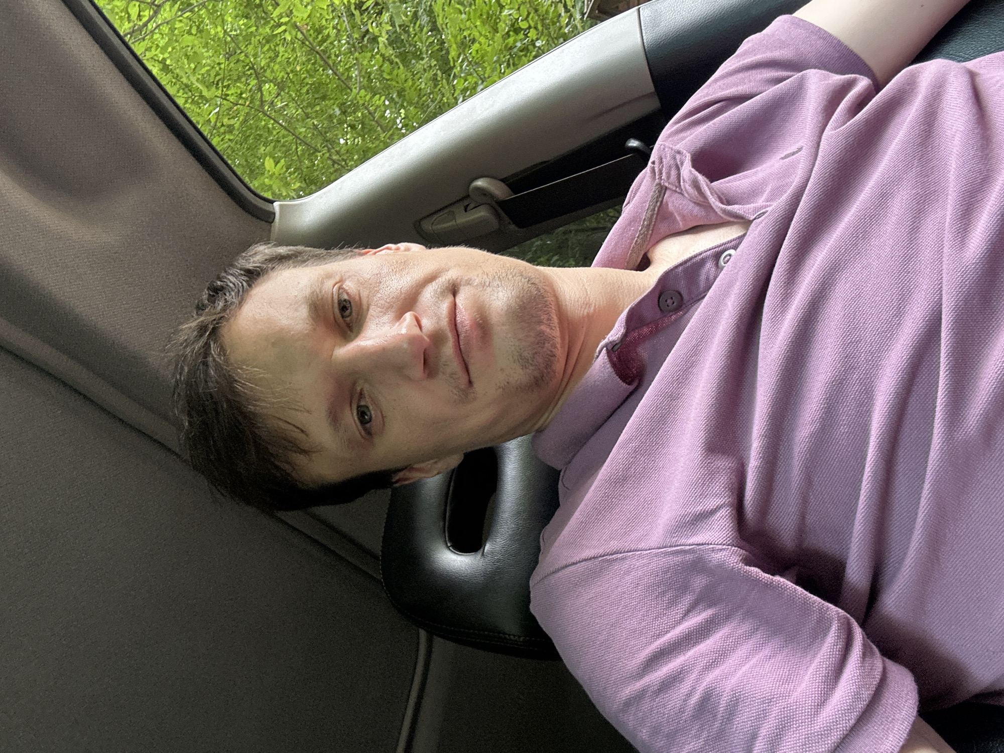 A man with short brown hair and light skin is sitting in the driver's seat of a car. He is wearing a light purple polo shirt. The interior of the car is visible, including the black headrest of the seat and the gray dashboard. Outside the car, green foliage and trees can be seen through the open window. The lighting suggests it is daytime. No specific location in Shatava, Ukraine, is identifiable from the image.