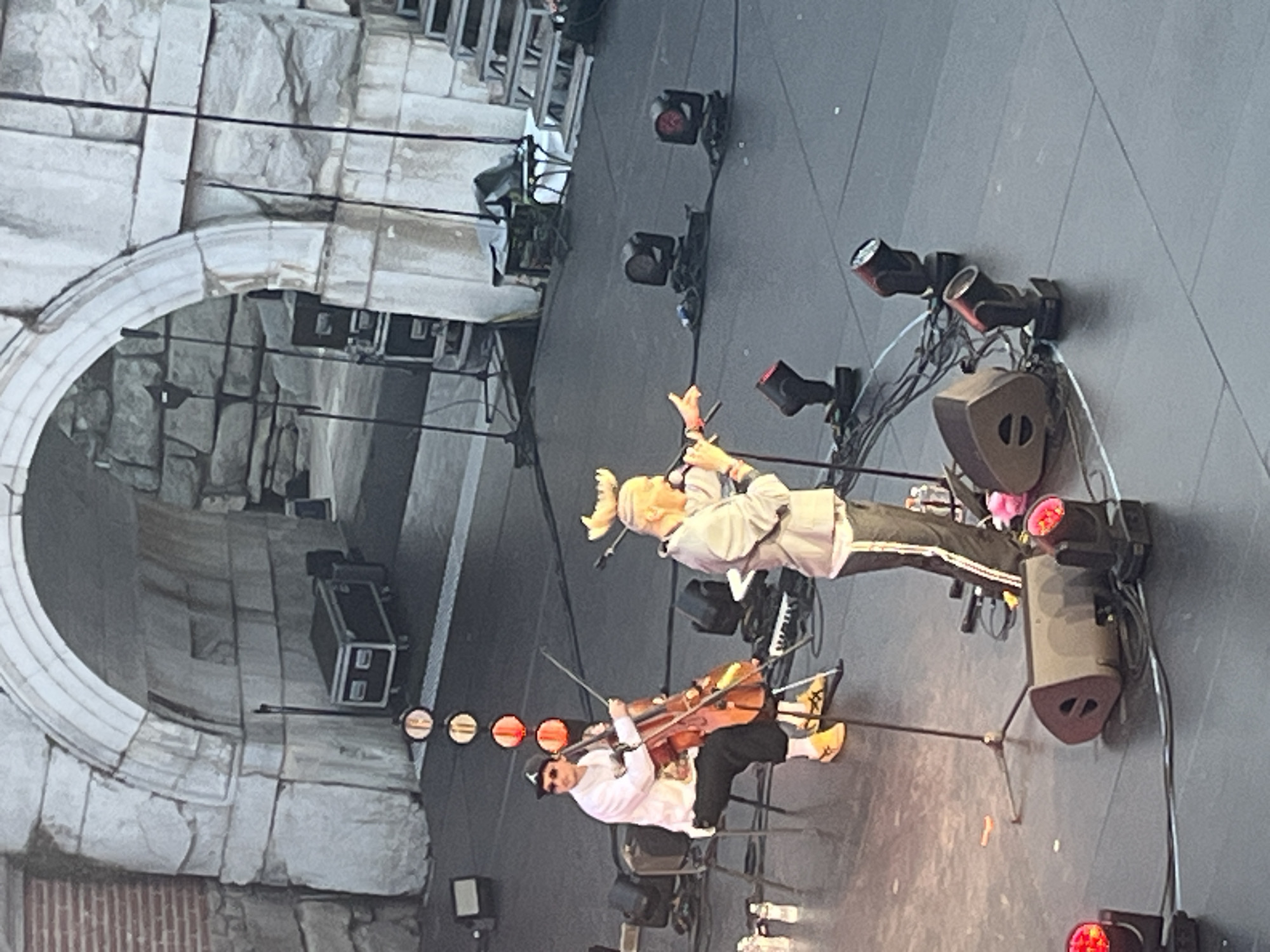 An outdoor concert stage is set in front of a large stone archway. Two male performers are on stage. The performer on the left is sitting on a chair, playing a cello. He is wearing a white long-sleeved shirt, black pants, a black baseball cap, sunglasses, and yellow sneakers. The performer on the right is standing and singing into a microphone. He is wearing a light gray jacket and dark pants with a white stripe. He has a novelty antler headband on his head. The stage floor is dark and tiled. There are several speakers and stage lights on the floor. In the background, through the archway, are large stone blocks and some equipment cases.