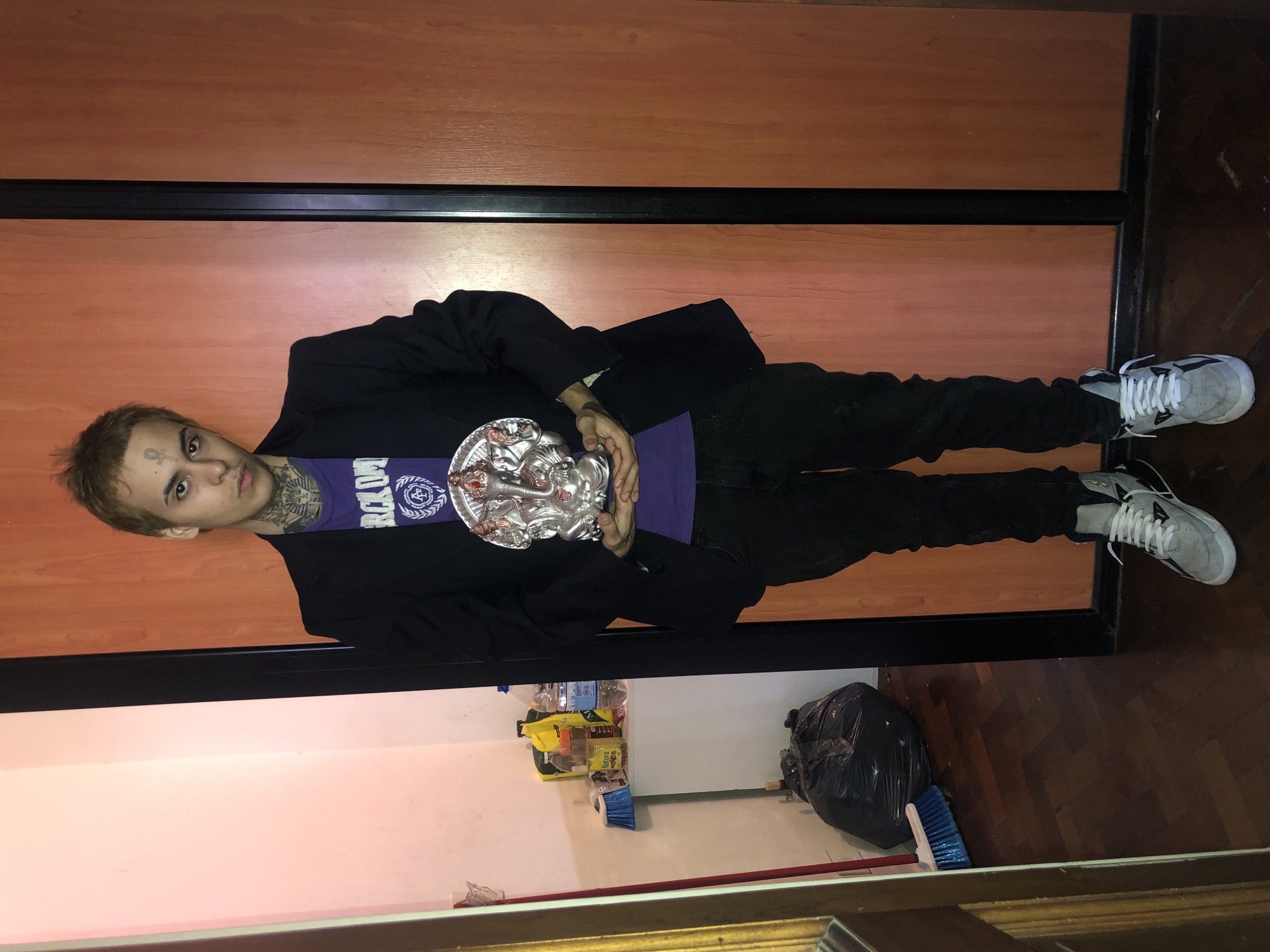 A young adult male stands indoors. He has light brown hair, tattoos on his face and neck, and is wearing a purple t-shirt with a white logo, a black blazer, black pants, and white sneakers. He holds a silver decorative object in his hands. Behind him is a wooden sliding door. In the lower portion of the frame, there are cleaning supplies, including a broom and a garbage bag. The wooden floor appears to be in Buenos Aires, Argentina.