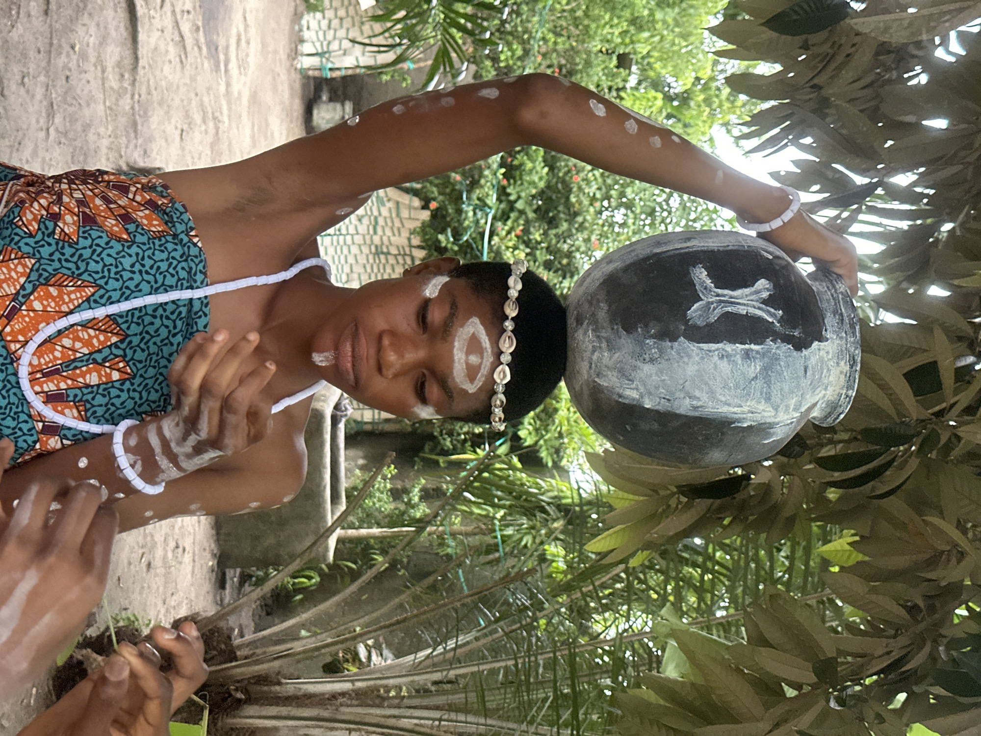 A young woman stands outdoors in Badagry, Nigeria, with a large, dark pot painted with white markings balanced on her head. She wears a beaded headband, a multi-colored patterned wrap dress, and white beaded necklaces and bracelets. White markings are painted on her forehead in an eye shape, on her chin, and dotted on her arms and shoulders. Her left arm is raised, supporting the pot, while her right hand is held in front of her, with another person's hands near it. The background features lush green foliage, including trees and palm fronds, and a concrete structure. The lighting suggests it is daytime, possibly overcast.