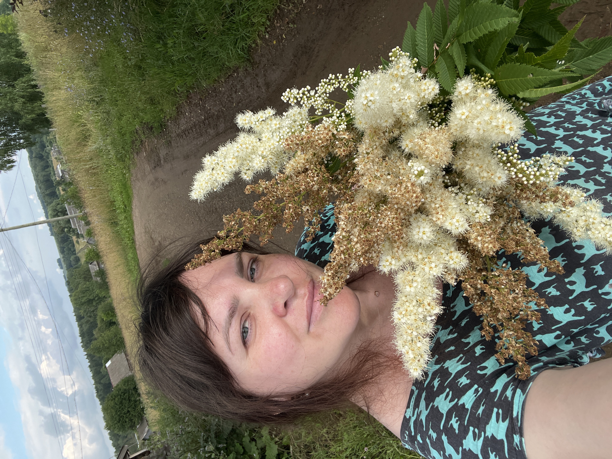 In Panteleevka, Russia, a woman with brown hair is captured in a selfie, holding a bouquet of white and brown flowers. She is wearing a dark shirt with a teal cat pattern. The background reveals a dirt road, a grassy field dotted with wildflowers, trees, and scattered buildings, suggesting a rural setting. The sky is partly cloudy.
