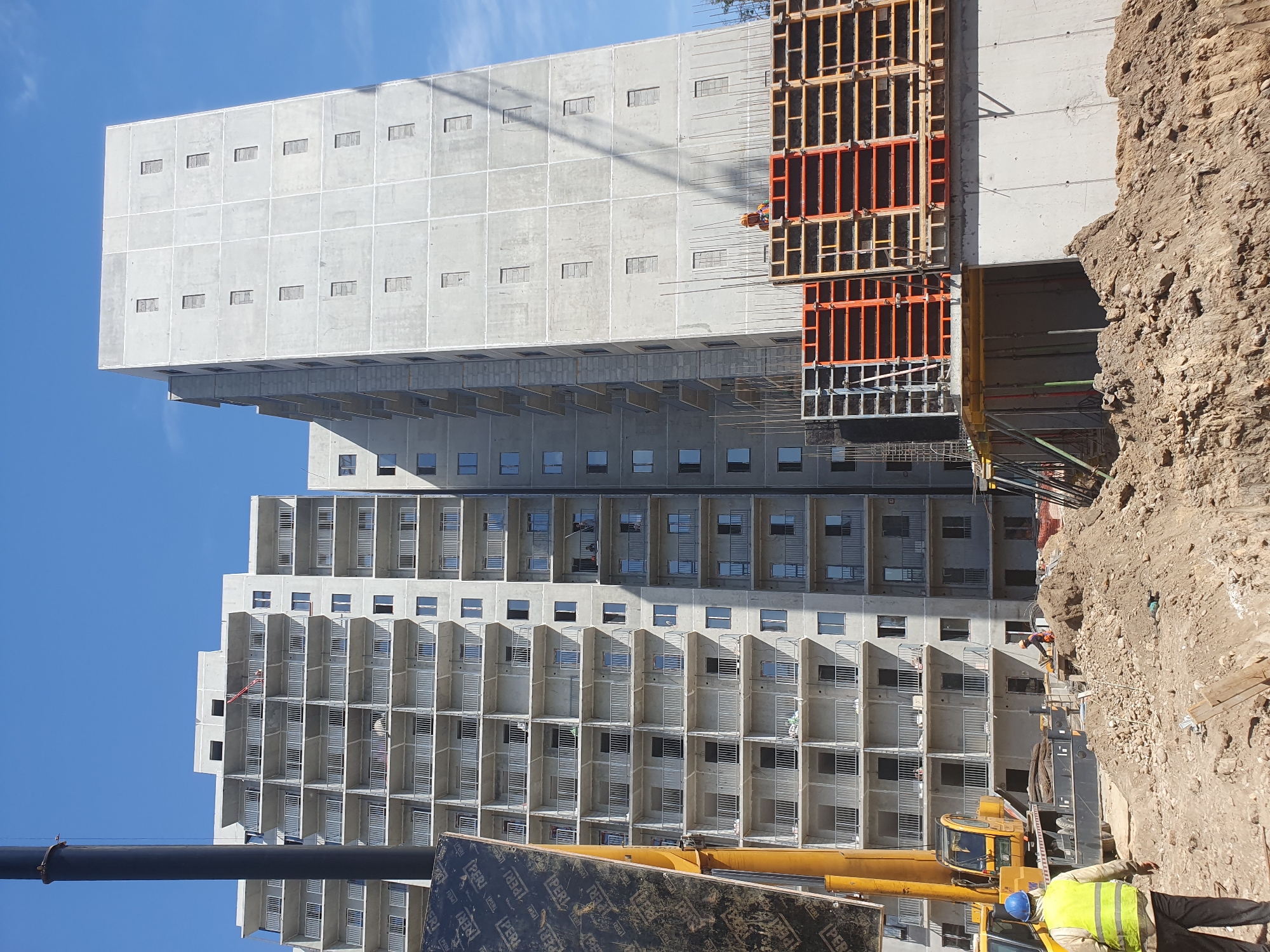 The image shows a construction site with two tall, unfinished apartment buildings in Tashkent, Uzbekistan. The buildings are made of concrete and have many balconies. One building is wider and taller than the other. A crane is visible on the left side of the image, and construction workers are present. The ground is covered in dirt and debris. The sky is clear and blue.
