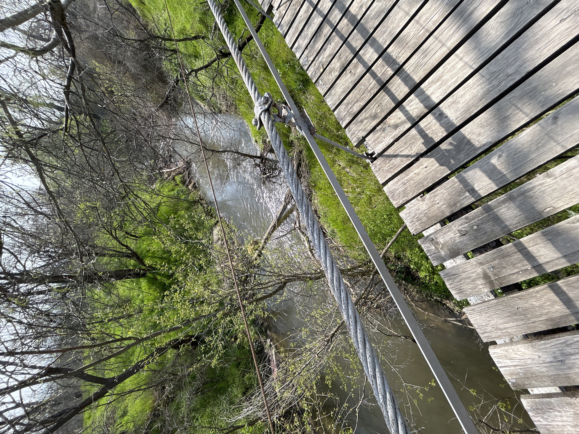 The image shows a section of a pedestrian suspension bridge over a stream. The bridge has wooden planks for the walkway, and thick steel cables support the bridge. The stream flows through a green area with trees. The image is taken from the bridge, looking down at the stream and the surrounding environment. There are no people visible in the image. Without additional information, it is not possible to confirm the location in the city of Plovdiv, Bulgaria.