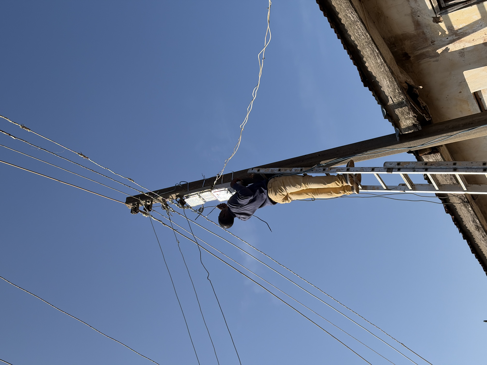 A man is working on a utility pole in Jos, Nigeria. He is standing on a ladder, reaching up to the wires and electrical equipment attached to the pole. The man is wearing a blue long-sleeved shirt and tan pants. Several electrical wires are visible, running across the frame. The sky is clear and blue. A portion of a building with a corrugated metal roof is visible in the lower right corner of the image.