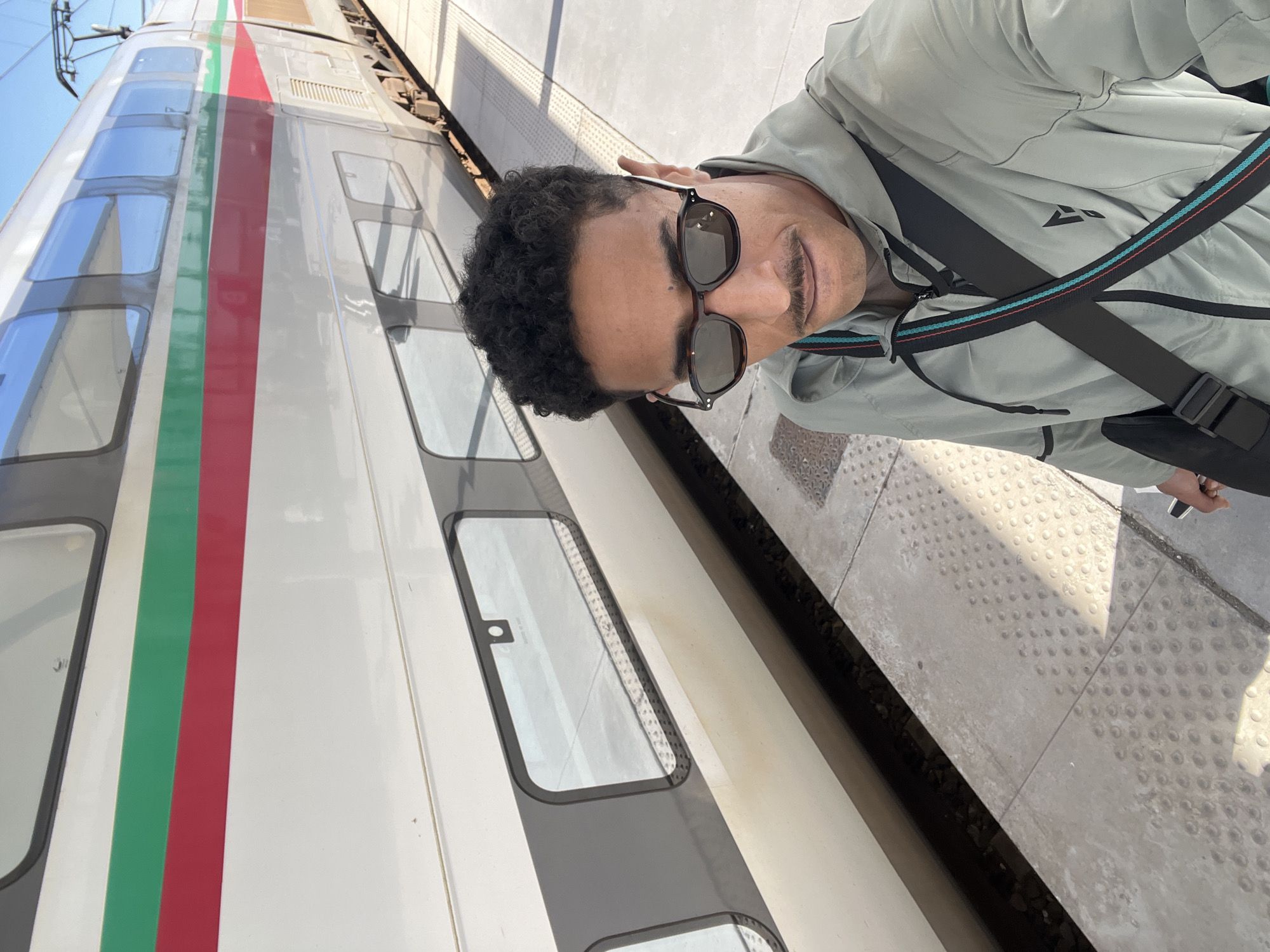 A male individual is captured in a selfie at the Tanger Ville railway station in Tanger, Morocco, standing on a light-colored concrete platform beside a stationary Al-Boraq high-speed train. The man, approximately 30-40 years old, has dark, curly hair, a thick mustache, and is wearing dark sunglasses with tortoiseshell frames. He is dressed in a light grey or sage green hooded jacket with a black shoulder bag featuring a strap with red and teal stripes. The train, a modern double-decker model, is predominantly white with horizontal red and green stripes running along its side. Sunlight casts distinct shadows on the platform, indicating clear weather. Overhead catenary lines are visible above the train in the background against a clear blue sky.