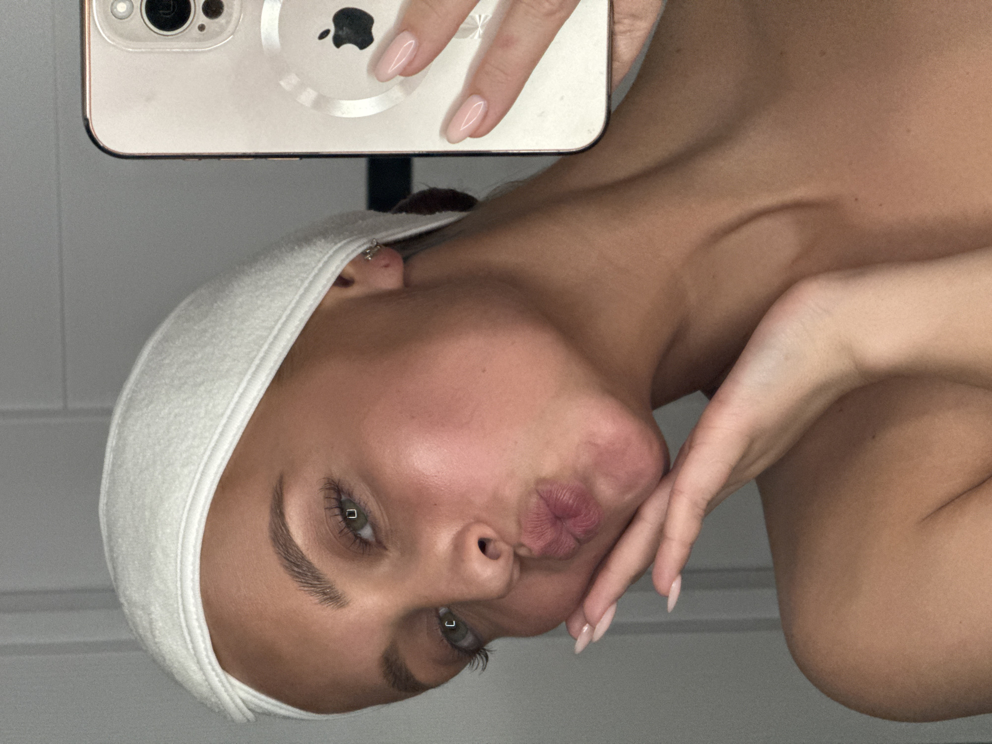 A woman with fair skin, green eyes, and light brown hair is taking a selfie in an indoor setting. She is wearing a white headband and has light pink nail polish. She is holding a smartphone with a visible Apple logo in her right hand. Her lips are pursed in a kissing gesture. The background appears to be a plain wall, possibly in a residential area. No specific information is present to suggest that the image was taken in Chaikovsky, Russia.