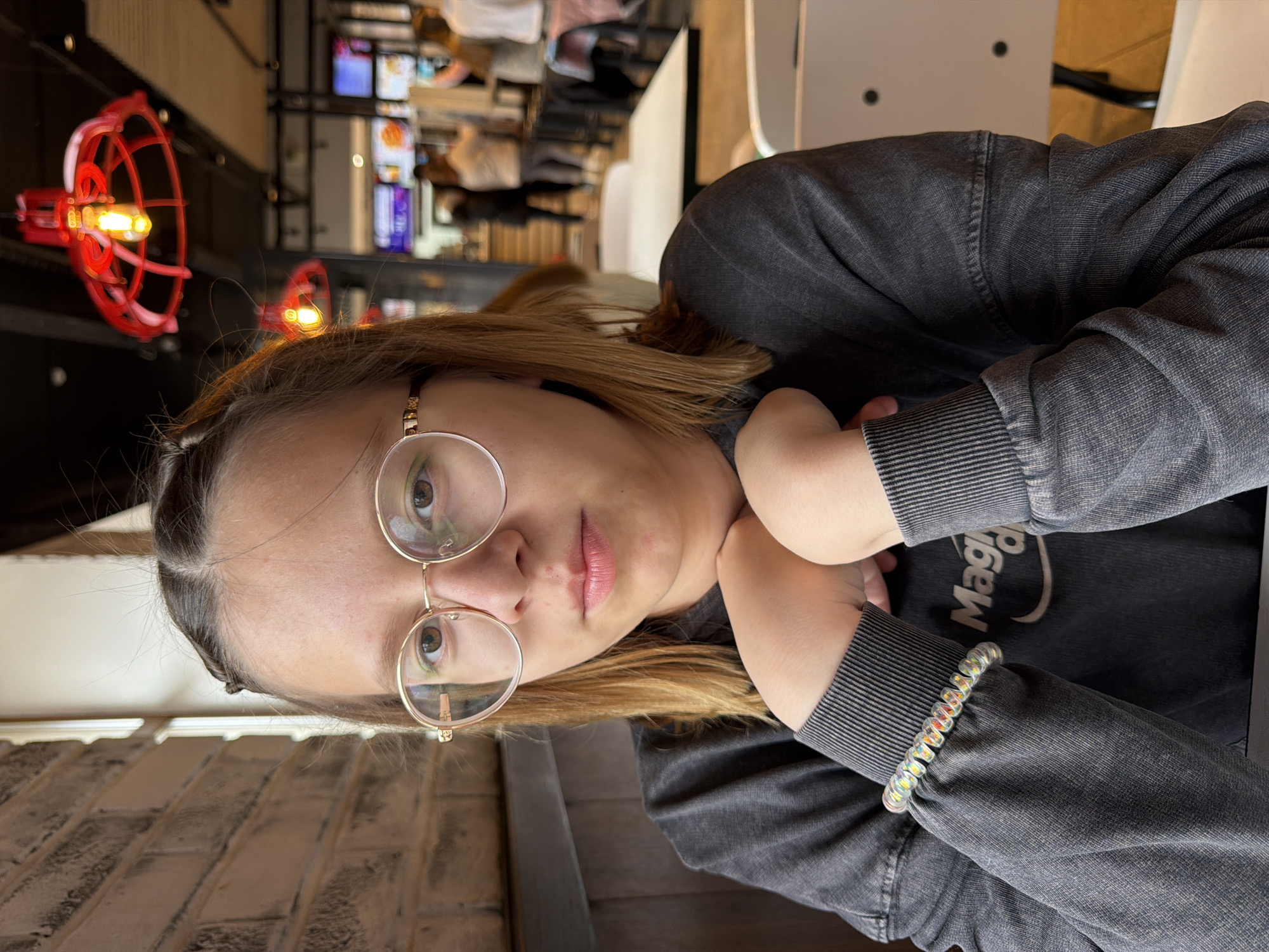 A young woman with blond hair pulled back in a partial braid and wearing round gold-framed glasses, a gray sweatshirt, and a clear coil bracelet is sitting at a table in what appears to be a restaurant. She is resting her chin on her hands and looking at the camera.
Behind her, out of focus, is a restaurant interior with other patrons and industrial-style lighting fixtures consisting of red wire cages around light bulbs. The location is in the city of Chaikovsky, Russia.