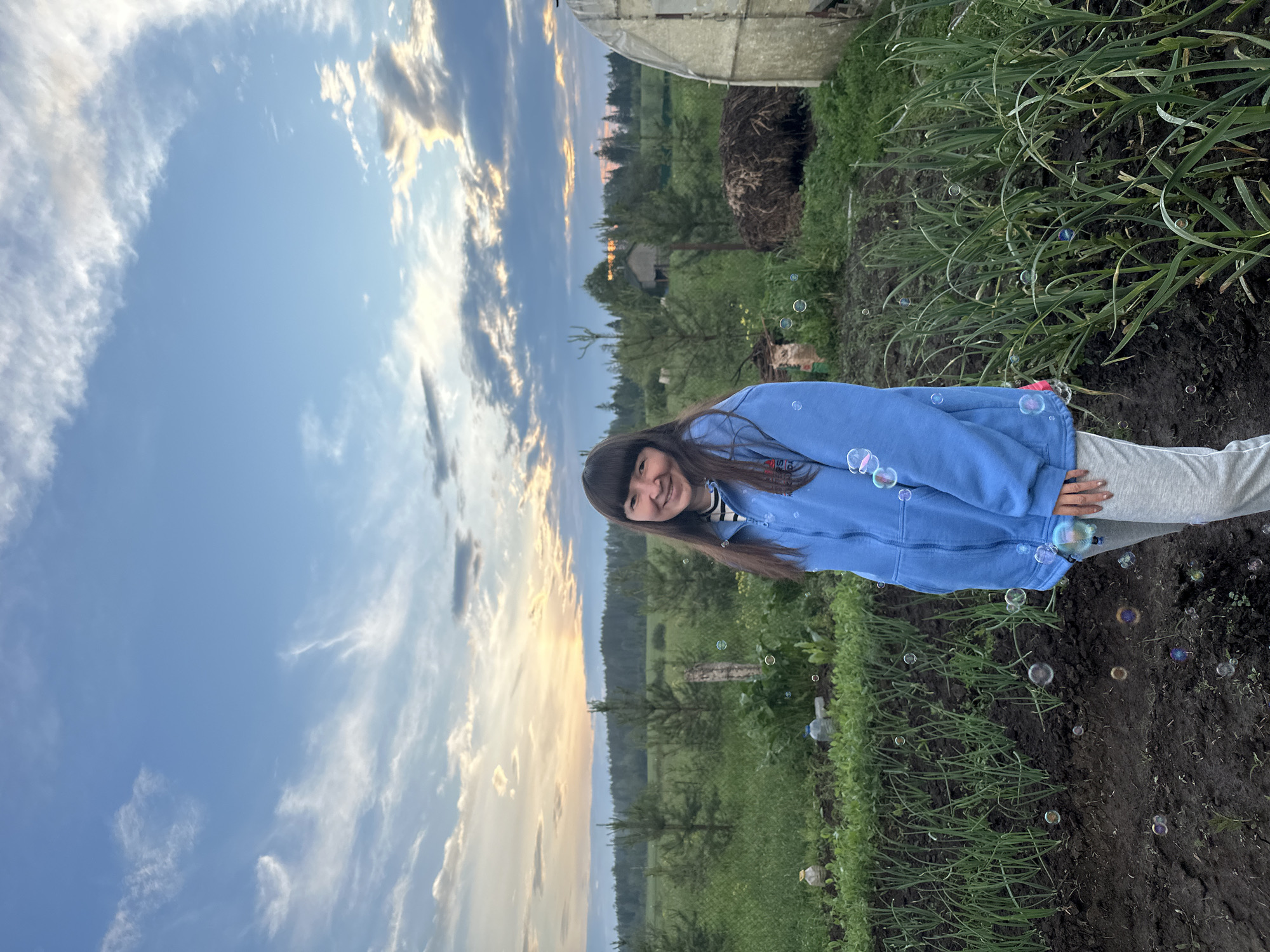 A young woman with dark hair and bangs, wearing a blue hoodie and grey sweatpants, stands in a garden. She is smiling and looking at the camera. In the background, there are rows of green plants, scattered trees, a small building, and a greenhouse. The sky is blue with scattered clouds, and the sun appears to be setting. Several soap bubbles are floating in the air around the woman and in the garden. The location is identified as Buranovo, Russia.