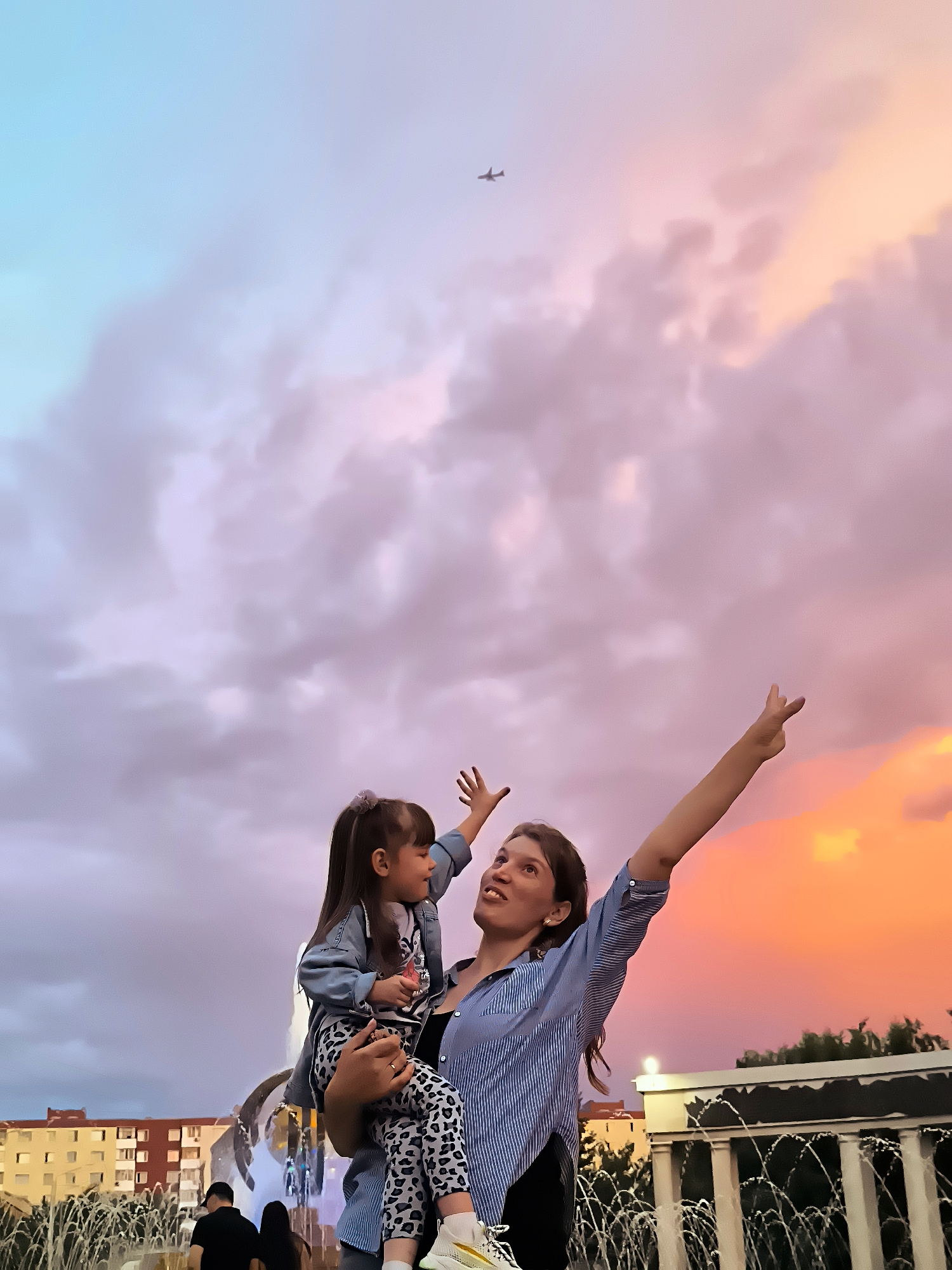 A woman holds a young girl in her arms, both looking up at the sky. The woman has her arms outstretched, and the girl is looking towards the sky with a smile. The background features a colorful sunset with hues of pink, orange, and blue. There is a fountain in the foreground and buildings in the distance, suggesting an urban setting. The sky also shows a small airplane in flight. The overall mood is joyful and celebratory. The setting appears to be Qarağandı city administration, Kazakhstan, given the context. The time of day is sunset.
