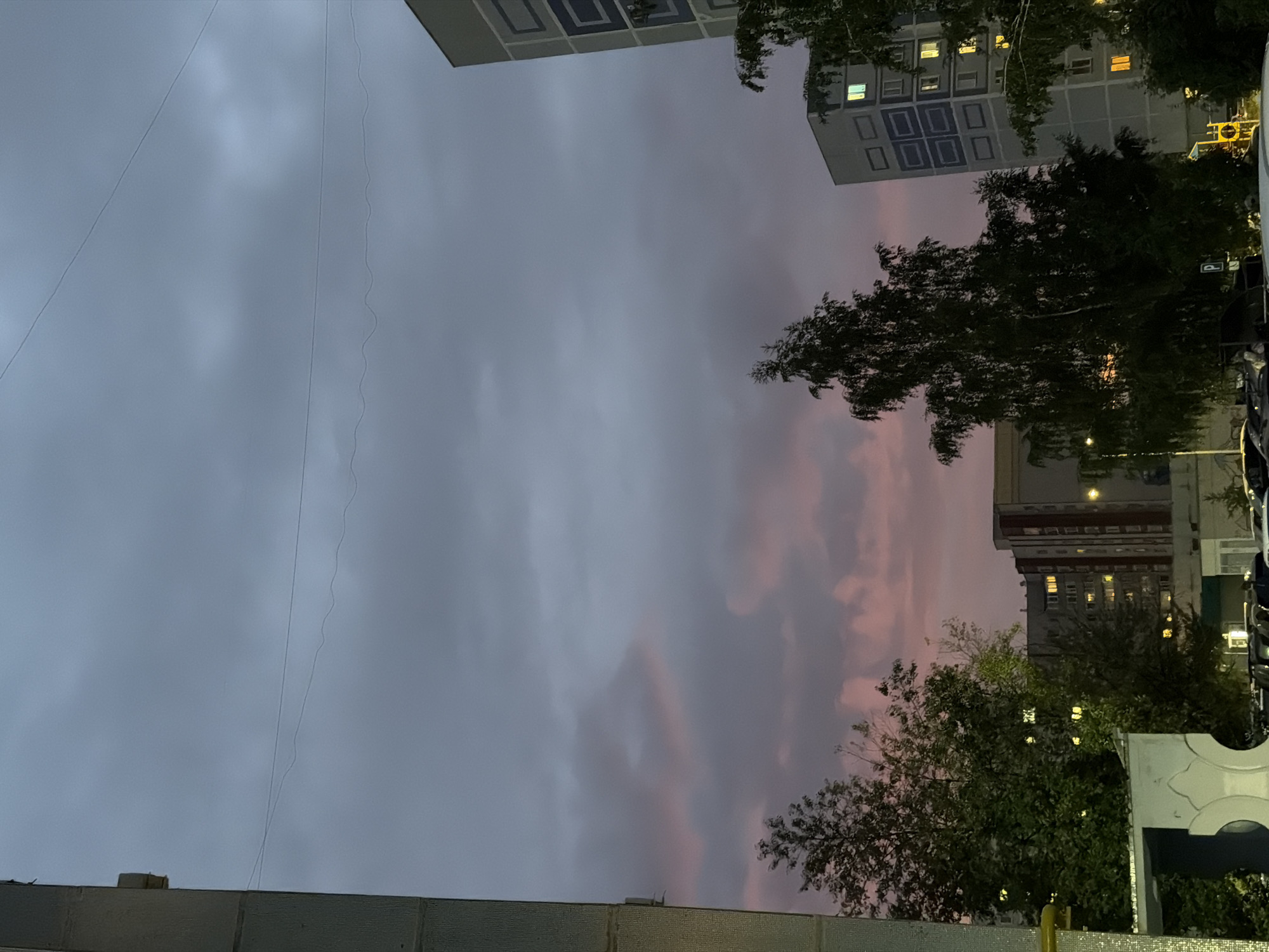 This media file captures a twilight scene with buildings, trees, and a cloudy sky. The composition is oriented vertically, with the sky occupying the majority of the frame. The sky displays a gradient of colors, transitioning from a muted gray on the left to soft pink and purple hues on the right, suggesting sunset or sunrise. Wispy clouds are scattered throughout, with some appearing denser and more illuminated by the fading light.
In the upper portion of the image, the facades of apartment buildings are visible. One building on the left, with a lighter color, features a grid-like pattern of dark rectangles. Another building on the right, darker in tone, has numerous windows illuminated with warm yellow light, indicating that it is inhabited. The perspective is looking upwards, creating a sense of height and scale.
Green trees with dense foliage are interspersed between the buildings and the sky. Their branches are silhouetted against the sky, with some leaves appearing illuminated by the ambient light. The overall atmosphere is calm and perhaps a little melancholic, typical of twilight in an urban environment.
At the bottom of the frame, a portion of a building's roof or exterior wall is visible, featuring a textured surface. A thin, wavy line, possibly a wire or string, is suspended in the sky, adding a subtle detail to the scene. There is no visible text in the media file. The time of day is clearly twilight, and the weather appears to be overcast with some atmospheric color. The location cues, such as the architectural style of the buildings and the mention of Nizhnekamsk, Russia, place the scene in an urban residential area.