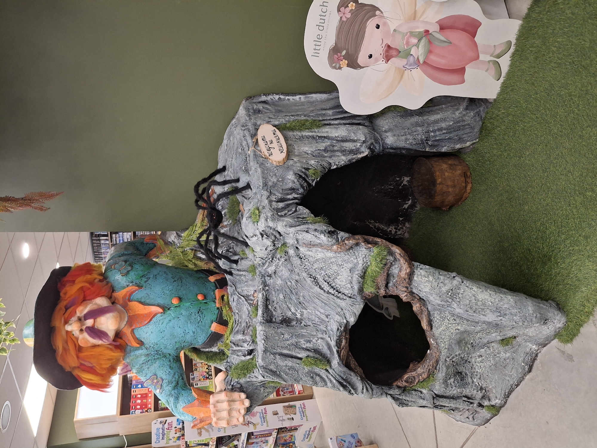 Here is a description of the image:
The image shows a whimsical, indoor display, likely within a retail environment such as a toy or children's bookstore.
The display features a large, gray, sculpted "cave" or rocky structure, possibly made of papier-mâché or a similar material. The structure includes several openings that suggest it could be a play area. There are faux spiders and moss-like details adding to the fantastical atmosphere. There's a sign "пещерата на желанията" attached to the rocks.
To the left of the structure, there's a colorful, whimsical figure of a character, perhaps a gnome or fairy-tale figure, with a black hat, orange hair, and a blue outfit.
Behind the cave structure, there is an artificial patch of green grass.
The backdrop is a plain green wall. Shelves filled with books and various children's products are visible.
A "little dutch" sign can be seen on the top right, attached to the rocky structure.
The location is indoors and seems to be a commercial space or a store.