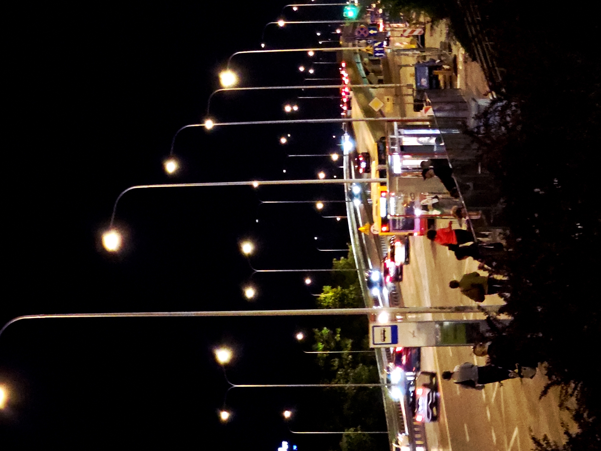 The image captures an urban street or bridge scene at night, viewed from an elevated perspective. The main subjects are numerous streetlights, vehicles, and several people on a sidewalk. The sky is dark black, contrasting sharply with the bright, warm-toned artificial lights that illuminate the roadway and surrounding areas.
Many streetlights with glowing, spherical bulbs extend into the distance along the left side of the frame, creating a strong sense of depth and leading lines. On the right, a multi-lane road is visible, populated by cars with illuminated headlights and taillights, suggesting traffic flow or vehicles stopped at an intersection. In the middle ground, several people are seen on a sidewalk or shoulder adjacent to the road. One person wears a bright red top, another a yellow or ochre top, and a third a light-colored top. They appear to be walking or standing.
Notable details include a green traffic light visible further down the road, as well as multiple red taillights. There are several construction barriers with orange and white stripes lining the road, indicating ongoing roadwork or a designated pathway. A bus stop sign, blue with a white "H" symbol, is clearly visible on a pole near the center of the right side. The scene is bustling with artificial light, creating a strong nocturnal atmosphere characteristic of a city environment.
