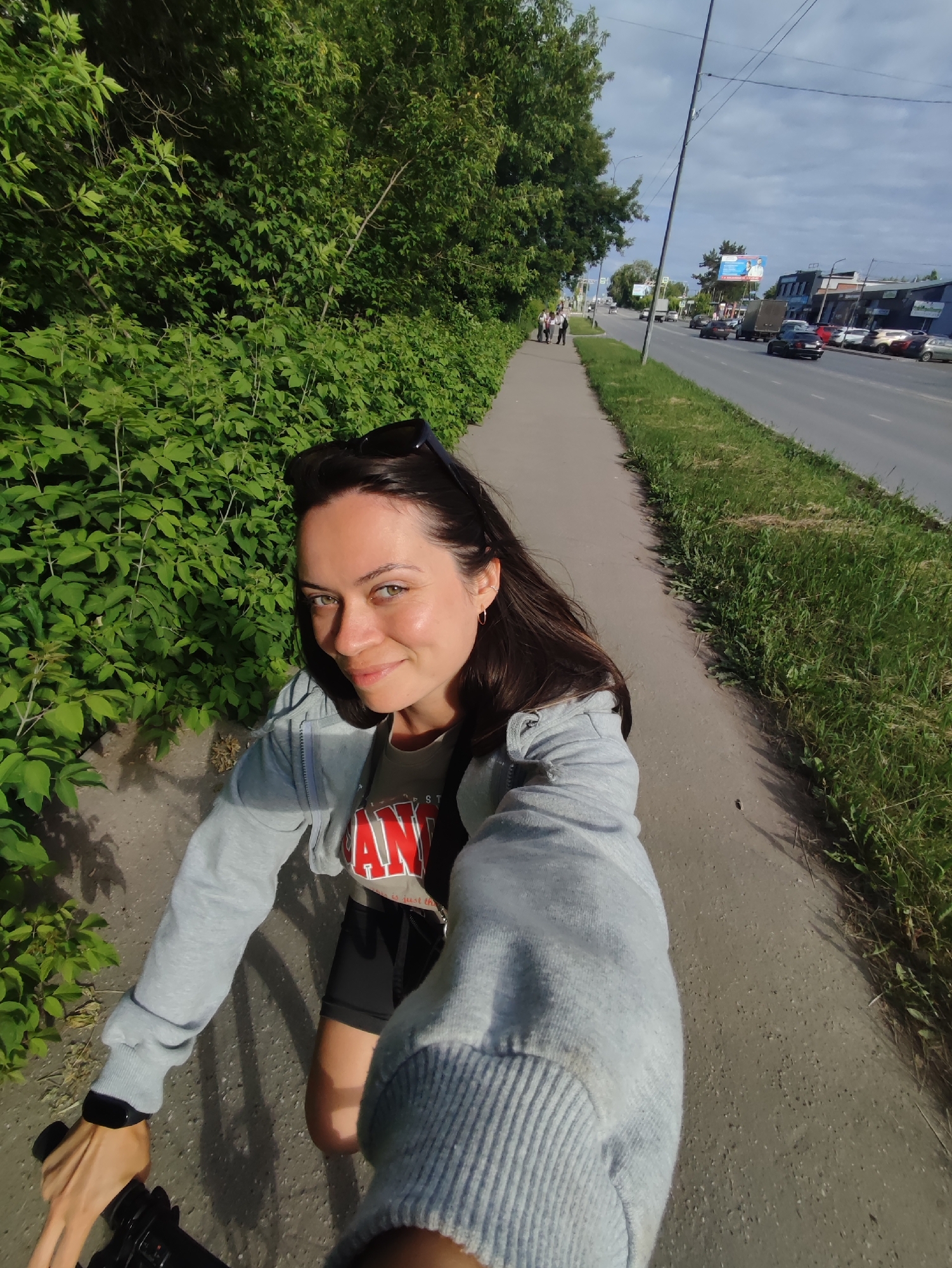 A woman is taking a selfie while cycling on a sidewalk in Chaikovsky, Russia. She is wearing a grey hooded jacket over a t-shirt and black shorts. Sunglasses are perched on her head. The sidewalk is next to a lush green hedge and a busy road with cars and trucks. In the background, buildings and billboards are visible, and the sky is partly cloudy. The overall scene depicts a sunny day in an urban environment.