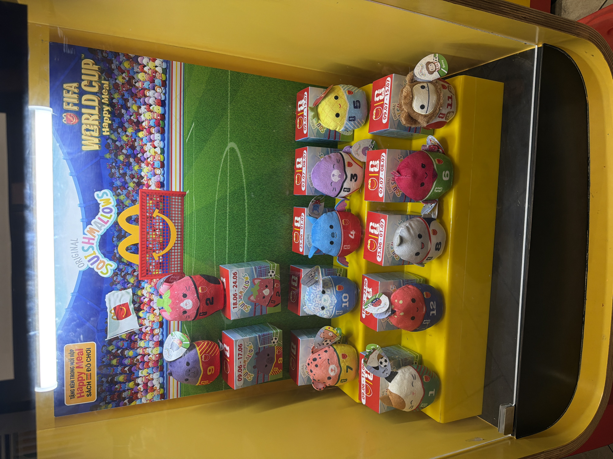 An indoor promotional display for McDonald's "FIFA World Cup Happy Meal" in Nha Trang Ward, Vietnam, features a yellow plastic case containing several rows of plush toys dressed in soccer-themed outfits. The backdrop depicts a stadium crowd and a soccer field. The toys are arranged on yellow shelves.
In the top row, two larger plush toys are visible: a purple bear wearing a red and yellow soccer jersey with the number 9 and a pink strawberry with a green stem wearing a red jersey with the number 2. Both are positioned above cardboard boxes.
The middle row displays six smaller plush toys. From left to right, they are: a pink leopard-print character in a yellow and red jersey with the number 7; a blue character in a blue jersey with the number 10; a light blue winged character in a red jersey with the number 4; a purple sea creature in a purple jersey with the number 3; and a yellow pineapple character in a blue and yellow jersey with the number 5.
The bottom row also features six plush toys. From left to right, they are: a white and brown cat character in a green and white jersey with the number 1; a red strawberry-like character in a blue and green jersey with the number 12; a grey round character in a white and blue jersey with the number 8; a pink strawberry character in a green and blue jersey with the number 6; and a brown monkey-like character in a red and white jersey with the number 11, identified as "Benny" on its tag.
The display is illuminated by overhead lights, and the overall environment appears to be a retail space.