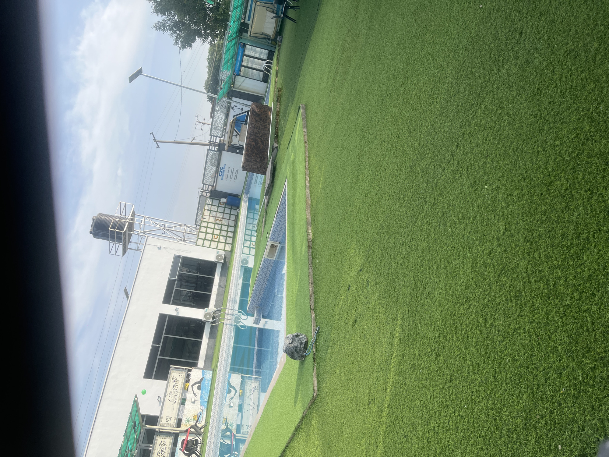 The image shows an outdoor swimming pool area with artificial turf. The pool has blue and white tiled accents. A large, dark grey rock sits on the edge of the turf next to the pool. Beyond the pool area, there is a white building with large windows. A water tank on a metal structure is visible above the building.
The scene is brightly lit, suggesting it is daytime with a clear or partly cloudy sky. Trees and some low-lying structures, possibly covered with green sheeting, are visible in the background. There are some power lines and a lamppost in the distance, indicating an urban or semi-urban setting.
One of the buildings has visible text: "G&C SWIMMING POOL" is written on a white rectangular sign. Below this, smaller text reads "YOU DON'T TRICK A". Further text is illegible. There are also some decorative murals or tiles on the side of the pool area depicting stylized human figures. The overall impression is a well-maintained recreational area. No people are visible in the image.