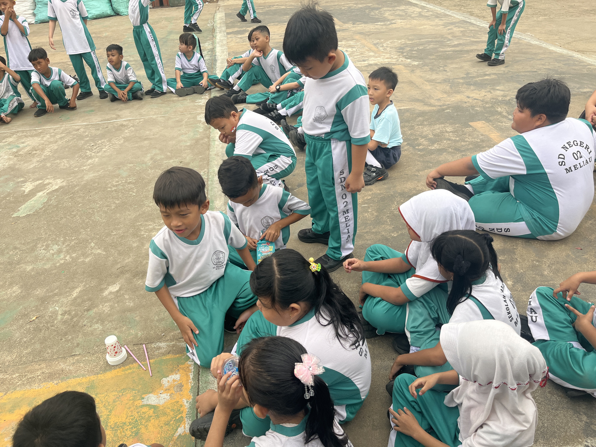 In Meliau Hulu, Indonesia, a group of schoolchildren in white and green uniforms are sitting and standing on a paved outdoor area. Some children are crouching on the ground, others are sitting cross-legged, and a few are standing. They appear to be engaged in some sort of activity or lesson, with some children looking at the ground or at each other. One child is holding a small bottle of water. Scattered on the ground are a few paper cups, a small stack of plates, and some pink straws. The environment suggests a schoolyard or a public space. The overall scene depicts a moment of organized activity for young students.