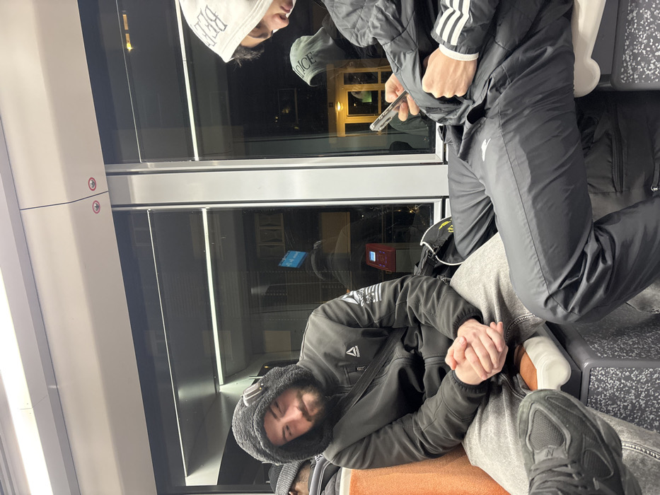 Two men are on a train at night. One man, wearing a black jacket and a hooded shirt, rests against the window. His eyes are closed, and his hands are clasped. The other man is standing beside him, facing away, wearing black Adidas track pants and jacket, and looking at his phone. The train's window shows reflections of buildings outside.
