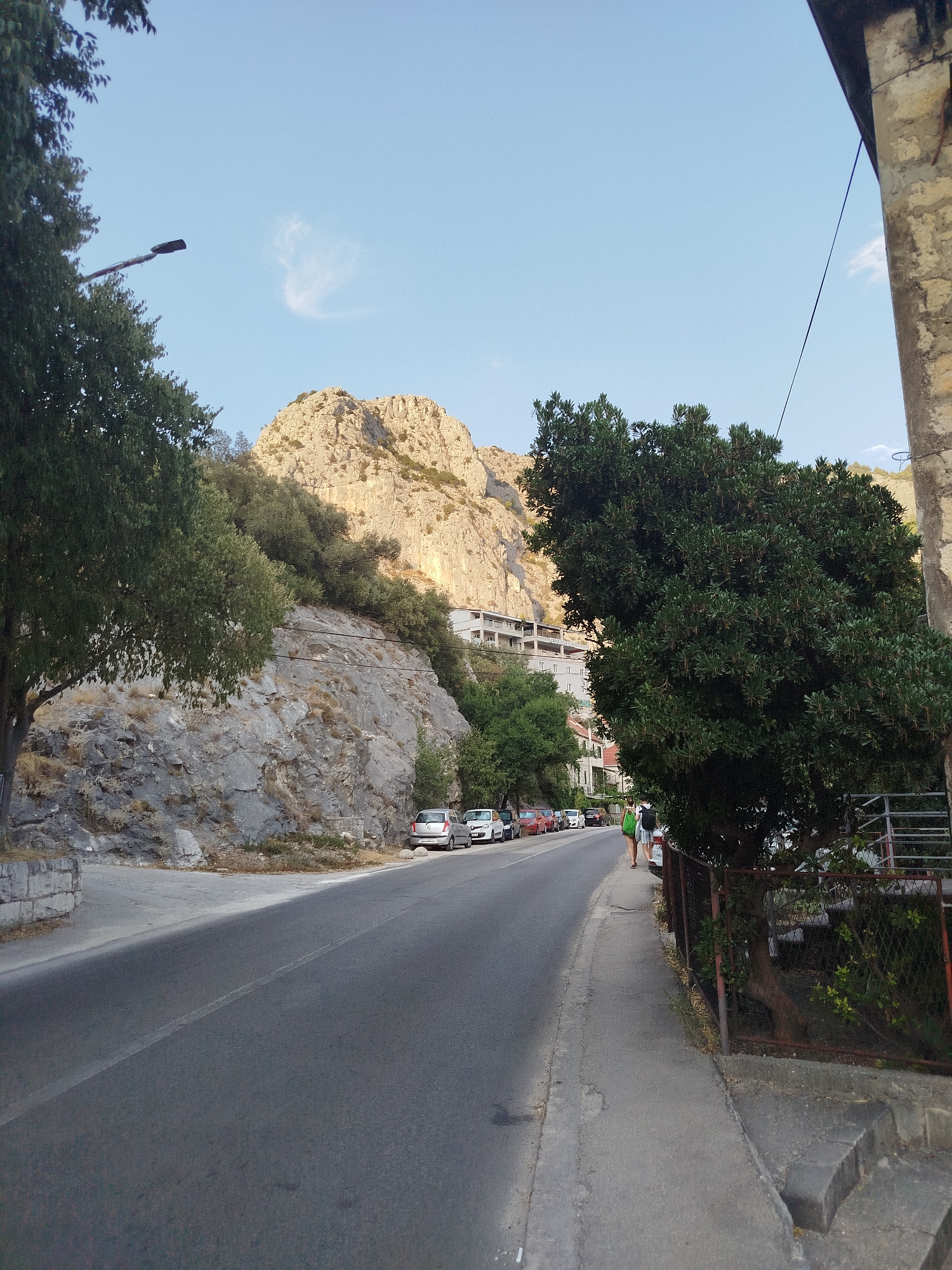 The image depicts a scenic road in Omiš, Croatia, winding alongside a rocky mountainside. The road is paved and has a white line marking the center. Several cars are parked along the left side of the road, and a sidewalk runs along the right. Two individuals are walking on the sidewalk, one carrying a backpack. Lush green trees flank the road and climb the rocky slopes. In the background, modern buildings are visible nestled against the mountain. The sky is a clear blue with a few wispy clouds. The overall impression is one of a picturesque coastal town nestled between the mountains and the sea.