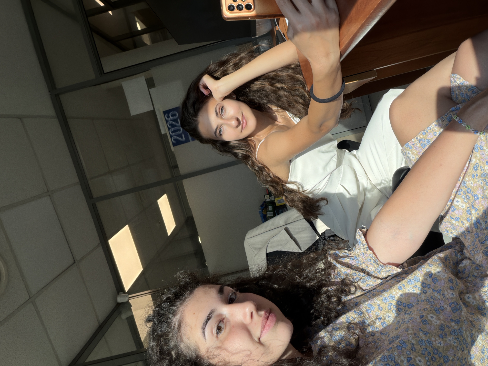 Two young women are seated in an office environment, likely in Sofia, Bulgaria. The woman on the left is in the foreground, facing the camera and smiling. She has long, curly dark hair and is wearing a light-colored dress with a floral pattern. The woman on the right is further back, looking towards the camera with her hand resting on her head. She has long, wavy dark hair and is wearing a white sleeveless top. She is holding a smartphone in her right hand, suggesting she might be taking a selfie or recording a video. The background features office partitions made of glass and metal, a ceiling with fluorescent lighting, and a sign on the glass wall displaying the number "2026". Sunlight is streaming into the room, casting shadows. A desk is visible in the foreground on the right, with a portion of a mobile phone on it. There are office chairs and some items on shelves in the background. The overall impression is a casual moment captured in a typical office setting.
