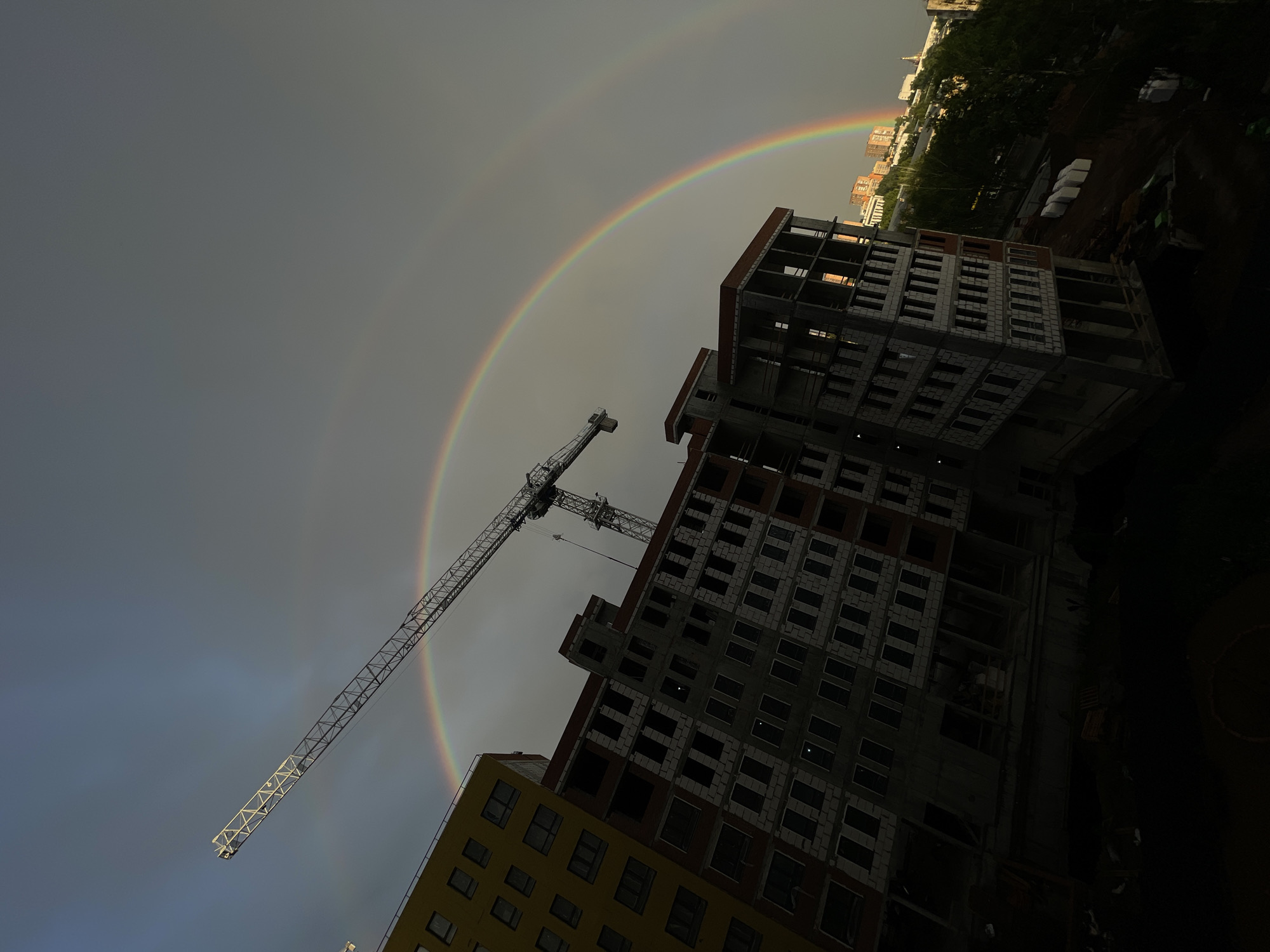 A double rainbow arches across a dark, overcast sky over a construction site in an urban environment. In the foreground, a yellow building with multiple windows is partially visible on the left, tilted at an angle. To its right, a multi-story concrete building under construction dominates the center of the frame, its windows dark and empty. A tall construction crane extends from the left side of the image into the sky, its boom angled upwards and to the right, intersecting with the primary rainbow. The secondary rainbow is fainter and appears above and parallel to the primary one. In the background, a cityscape with buildings and trees is visible, illuminated by the sunlight filtering through the clouds, creating a contrast with the darker foreground. The overall lighting suggests it is either early morning or late afternoon with a storm passing or imminent.