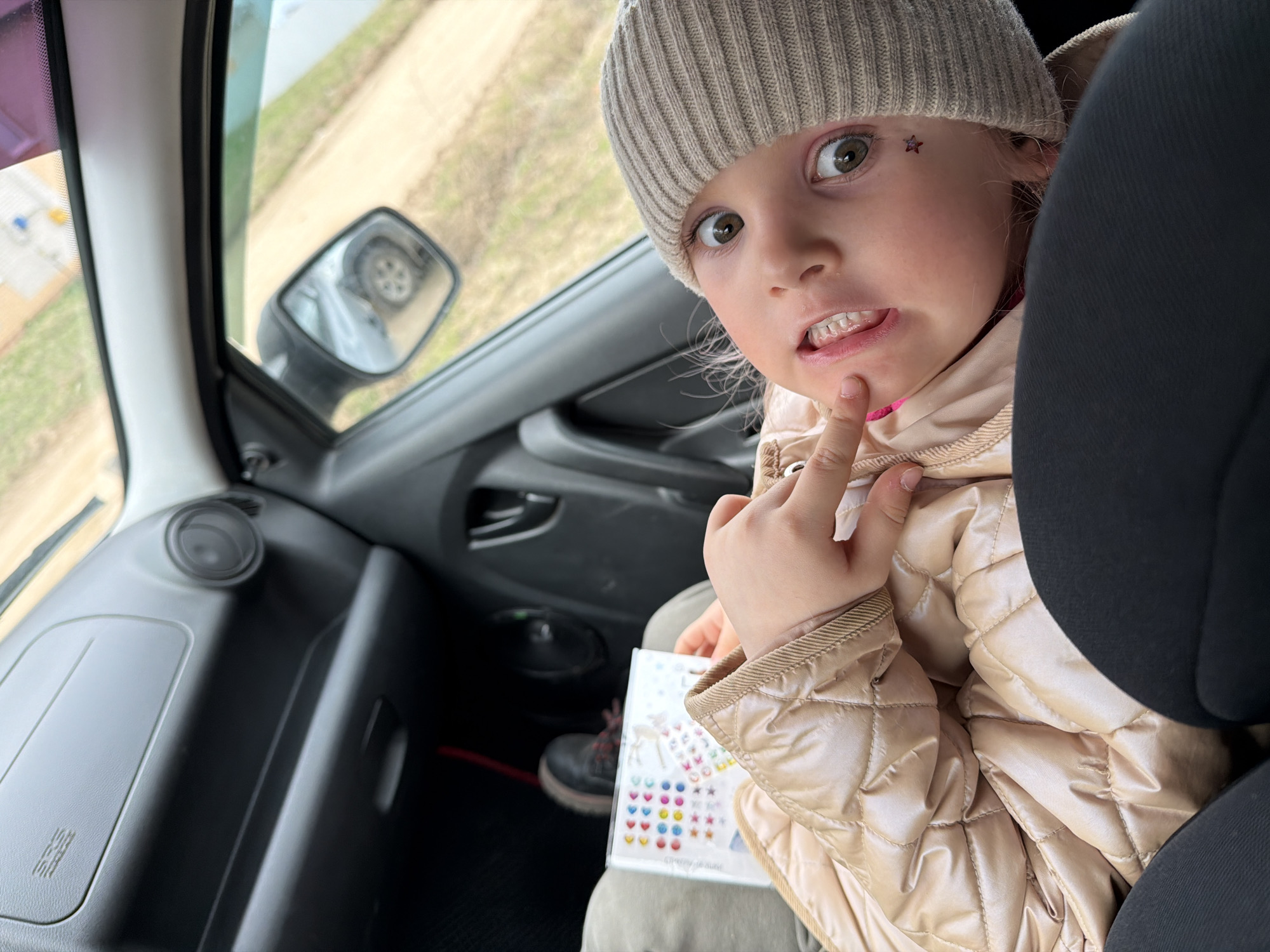 A child wearing a beige beanie and a light brown quilted jacket is sitting in a car. The child is looking at the camera and making a face with their hand on their chin. They have a star sticker on their forehead. The car's interior is visible, including the dashboard, side mirror, and part of a black seat. The child is holding a sticker sheet. The location is Chaikovsky, Russia, as the image metadata specifies this location.