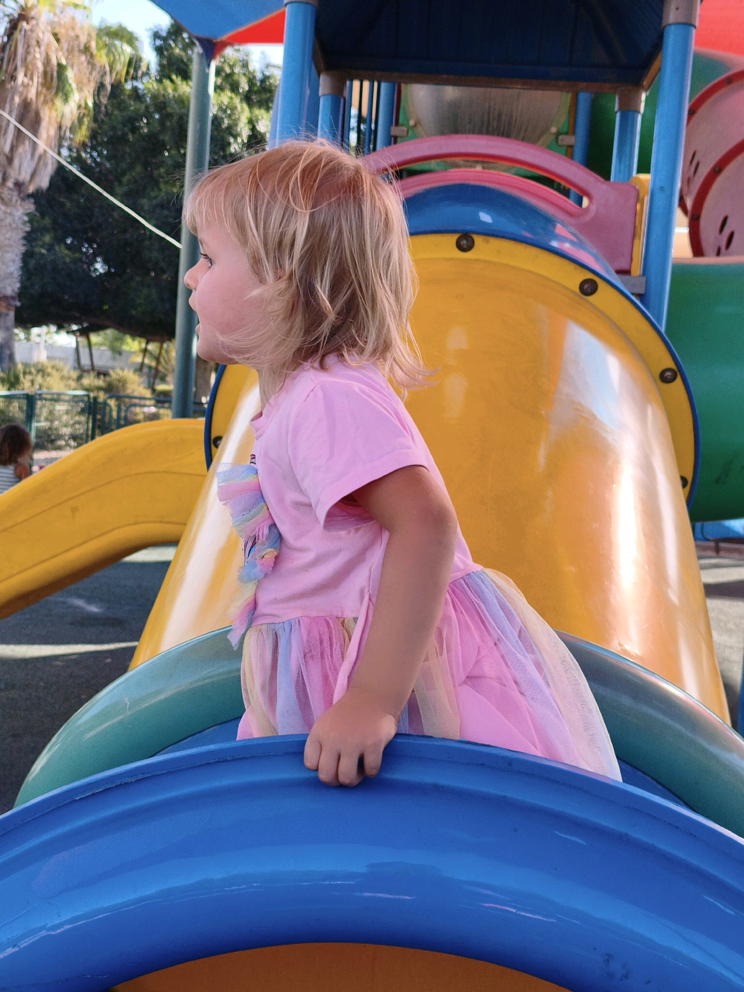 A young girl with blonde hair, wearing a pink t-shirt and a colorful tutu, is seen from the side at a playground. She is looking to her left. The playground features large, brightly colored plastic slides in yellow, blue, and green. The background shows trees and a fence. The image appears to have been taken outdoors on a sunny day. This scene is set in a playground within the Lachish Regional Council, Israel.