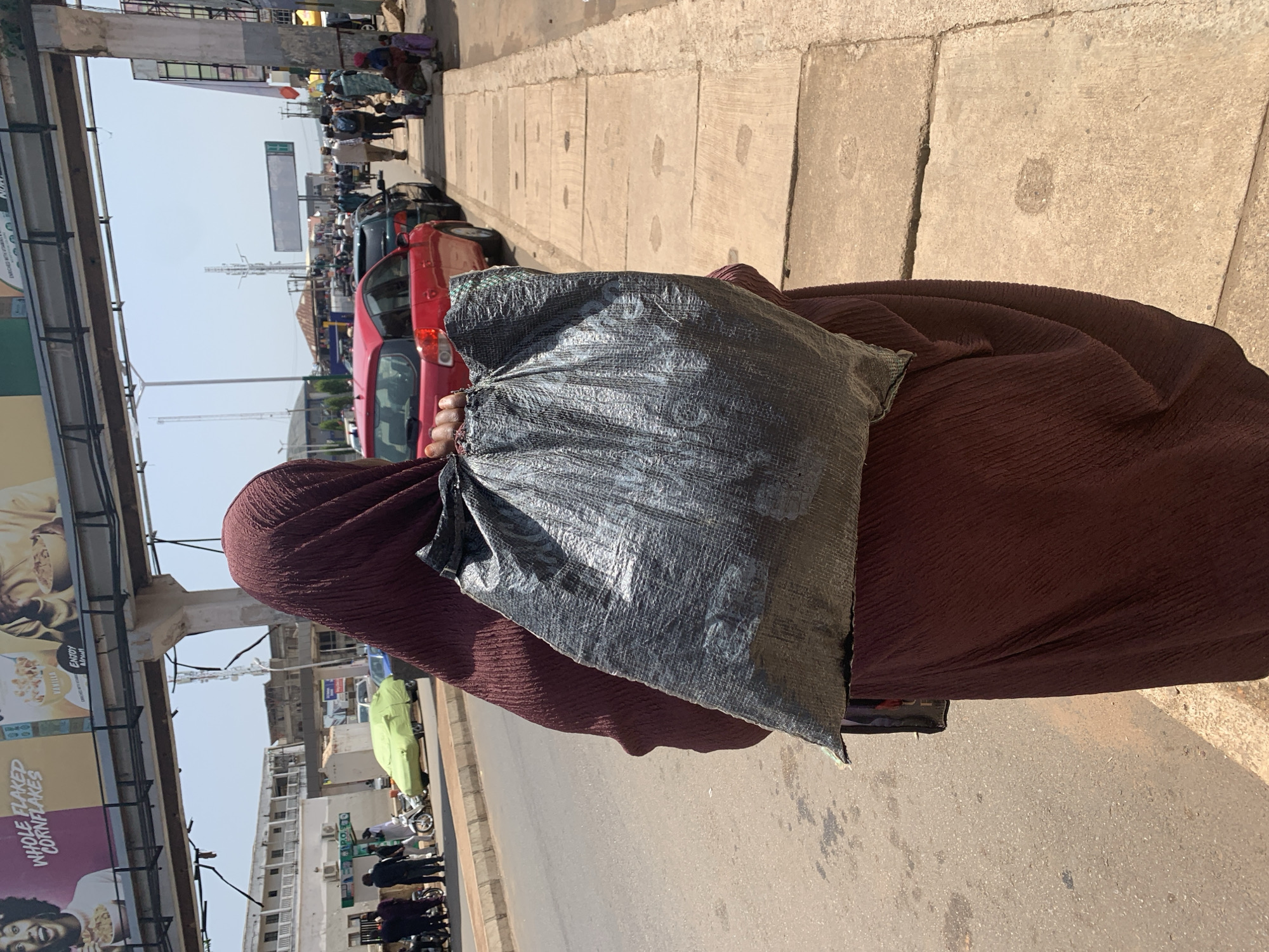 A person wearing a maroon hijab and dress carries a large, dark, crinkled plastic bag over their right shoulder. The bag appears to be filled and is held by the wearer's right hand. The person is walking on a paved sidewalk beside a street. On the street, a red car and other vehicles are visible. In the background, buildings and overhead infrastructure, possibly a bridge or flyover, can be seen. The scene appears to be in an urban environment, with street vendors and pedestrians also present further down the sidewalk. The overall impression is a busy street scene in Jos, Nigeria.