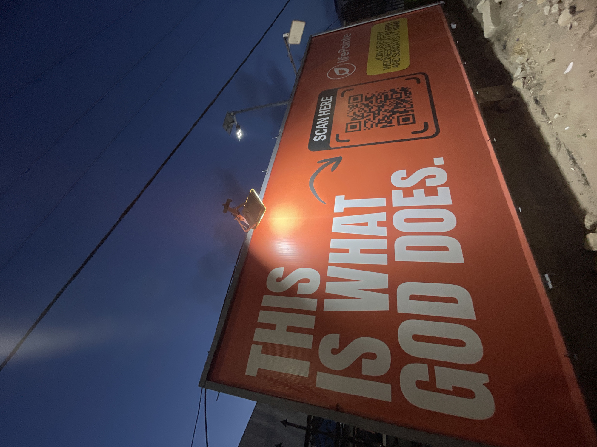 The image shows a sign for LifePointe Church in Ilado, Nigeria. The sign is red and prominently displays the text "THIS IS WHAT GOD DOES." It also features a QR code with the instruction "SCAN HERE," and mentions service times: "JOIN US EVERY WEDNESDAY AT 6:18PM AND SUNDAYS AT 10AM." The photo was taken at dusk. A utility pole and wires are also visible.