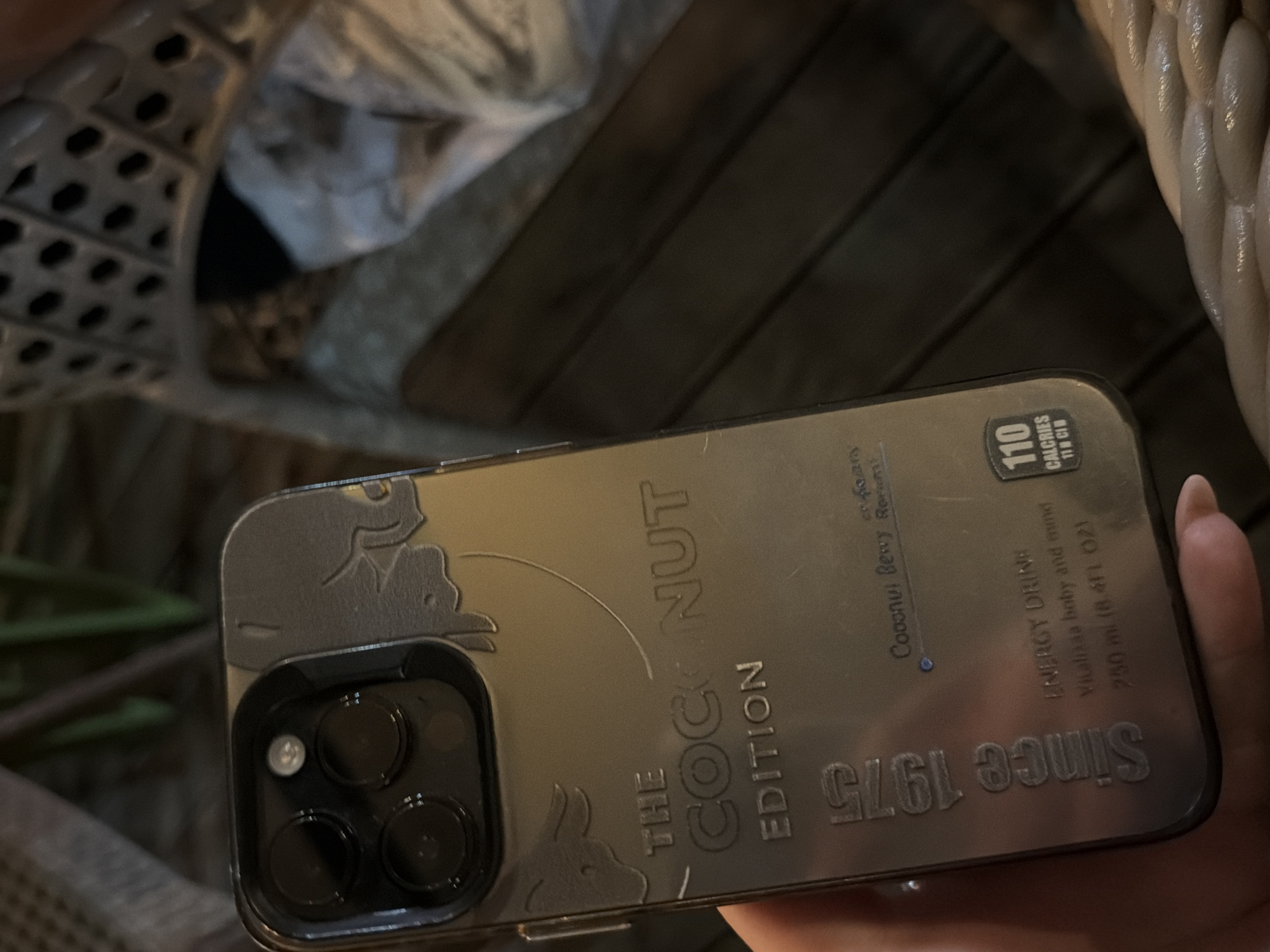 A smartphone with a decorative case resembling a vintage coconut beverage is held in a hand. The case features text "THE COCONUT EDITION", "Since 1975," and nutritional information, including "110 CALORIES". The phone has a multi-lens camera array. The background is out of focus, showing indistinct textures and patterns. No location details, specifically Elhovo, Bulgaria, are evident in the image.
