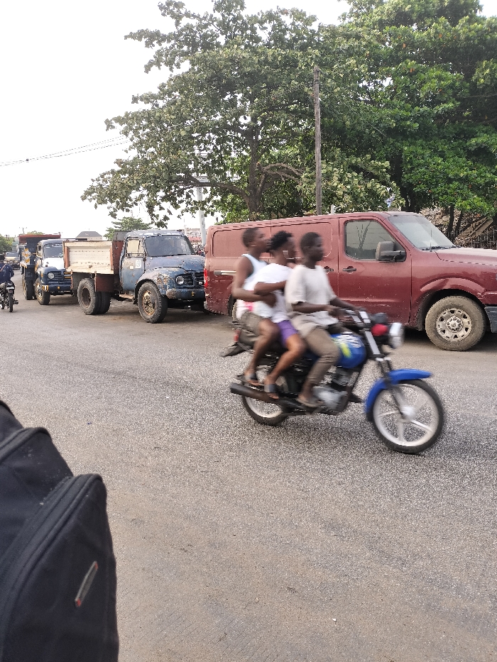 The image captures a dynamic street scene during daylight, likely in an urban or semi-urban setting, characterized by traffic and a mix of vehicles.
In the immediate foreground, a blurry black object, possibly a backpack or bag, occupies the lower-left corner, suggesting the photo was taken from a pedestrian's perspective or from another moving vehicle. The road surface visible is asphalt, showing some wear.
The central focus of the image is a blue motorcycle in motion, evidenced by the significant motion blur on its wheels and the three individuals riding it. The driver, positioned at the front, wears a light-colored t-shirt and what appears to be light brown or beige trousers. Immediately behind the driver is a person wearing a white top and purple shorts. The third person is seated at the very back, wearing a dark top and dark shorts. Their faces are indistinct due to the motion blur. The motorcycle itself is a standard blue model, possibly a commuter bike, with silver-colored rims.
To the right of the motorcycle, a dark red or maroon panel van is visible, parked or moving slowly. Its side door appears to be of the sliding variety.
Further into the midground, to the left of the motorcycle, a line of heavy-duty trucks stretches down the road. The most prominent is an older model blue truck with an open flatbed or dump bed, exhibiting visible signs of wear, dents, and rust, suggesting considerable use. Its tires appear robust and well-worn. Behind this blue truck, another truck of similar design, but a lighter color (possibly white or light blue), is also part of the queue. Further down the road, another smaller motorcycle with a rider is visible near the line of trucks.
The background is dominated by a dense canopy of lush green trees, particularly on the right side and across the upper portion of the frame, indicating a rich natural environment. A utility pole stands vertically among the trees on the right. The sky, visible in the upper-left, appears bright and hazy or overcast, typical of daylight conditions.
The overall scene conveys a sense of everyday life and traffic flow, with the overloaded motorcycle being a notable detail. No discernible text is visible on any of the vehicles or in the environment.