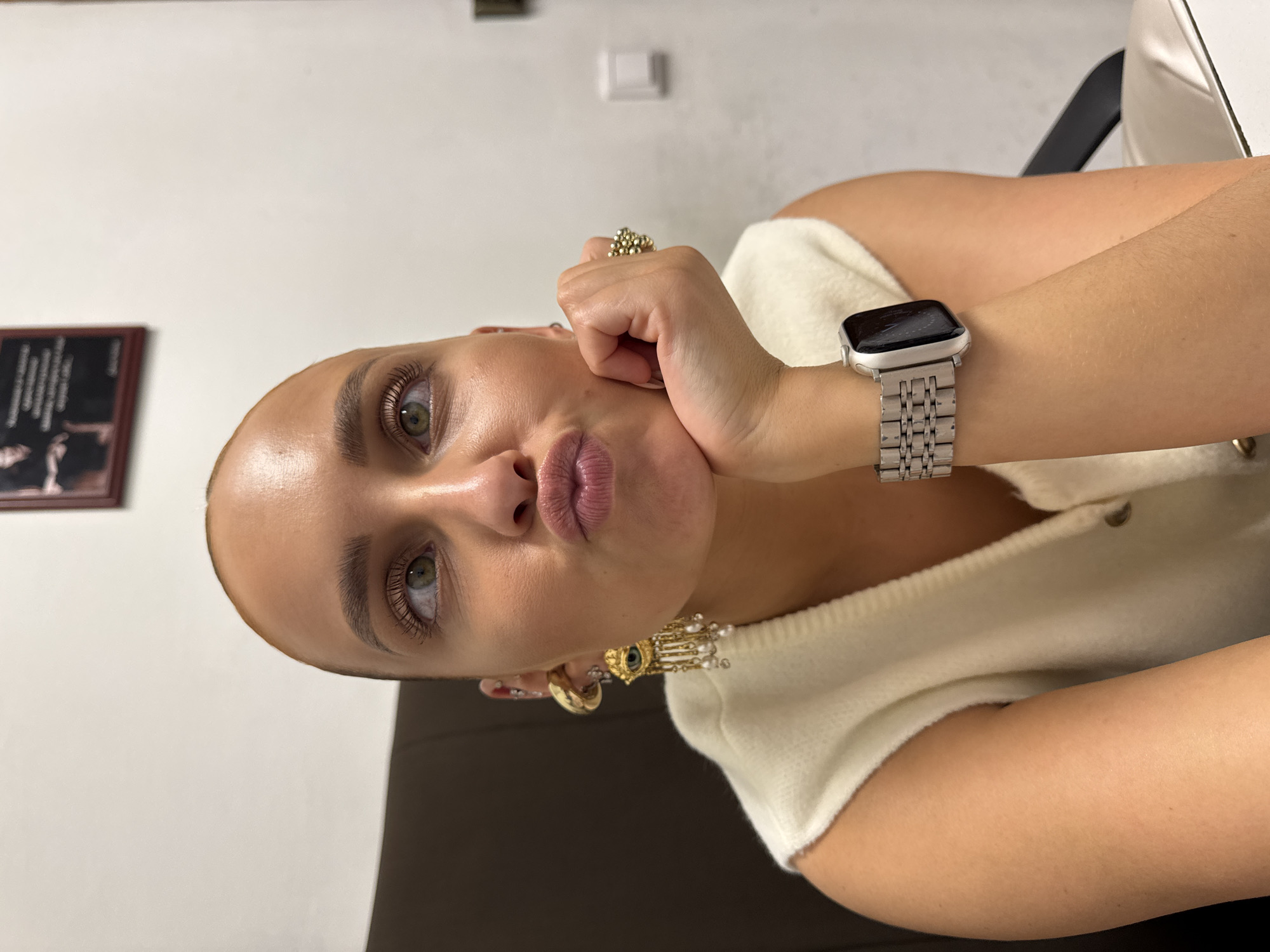 A female subject, appearing to be an adult, is depicted in a close-up portrait, looking upwards and to her left with pursed lips, resting her chin on her right hand. Her hair is extremely short and light-colored. She has prominent green or hazel eyes with visible eyelashes and well-groomed eyebrows. She wears a sleeveless, off-white, ribbed or knitted top with a V-neck and a single visible button closure. On her left ear, she has multiple piercings, including a large gold hoop earring and a dangling earring featuring an "evil eye" motif with blue and gold elements, from which multiple gold chains with small white pearls extend. On her right ring finger, she wears a cluster ring composed of numerous small gold-colored spheres. A silver-colored smartwatch with a metallic link band is visible on her left wrist, with a dark screen. The background consists primarily of a plain light-colored wall. A dark framed picture or document is partially visible on the upper right wall, and a white electrical wall switch is present on the lower right. A dark upholstered surface is visible behind her left shoulder, and a lighter-colored armrest or cushion is visible behind her right arm. The image does not contain identifiable elements to confirm the location as the city of Chaikovsky, Russia.