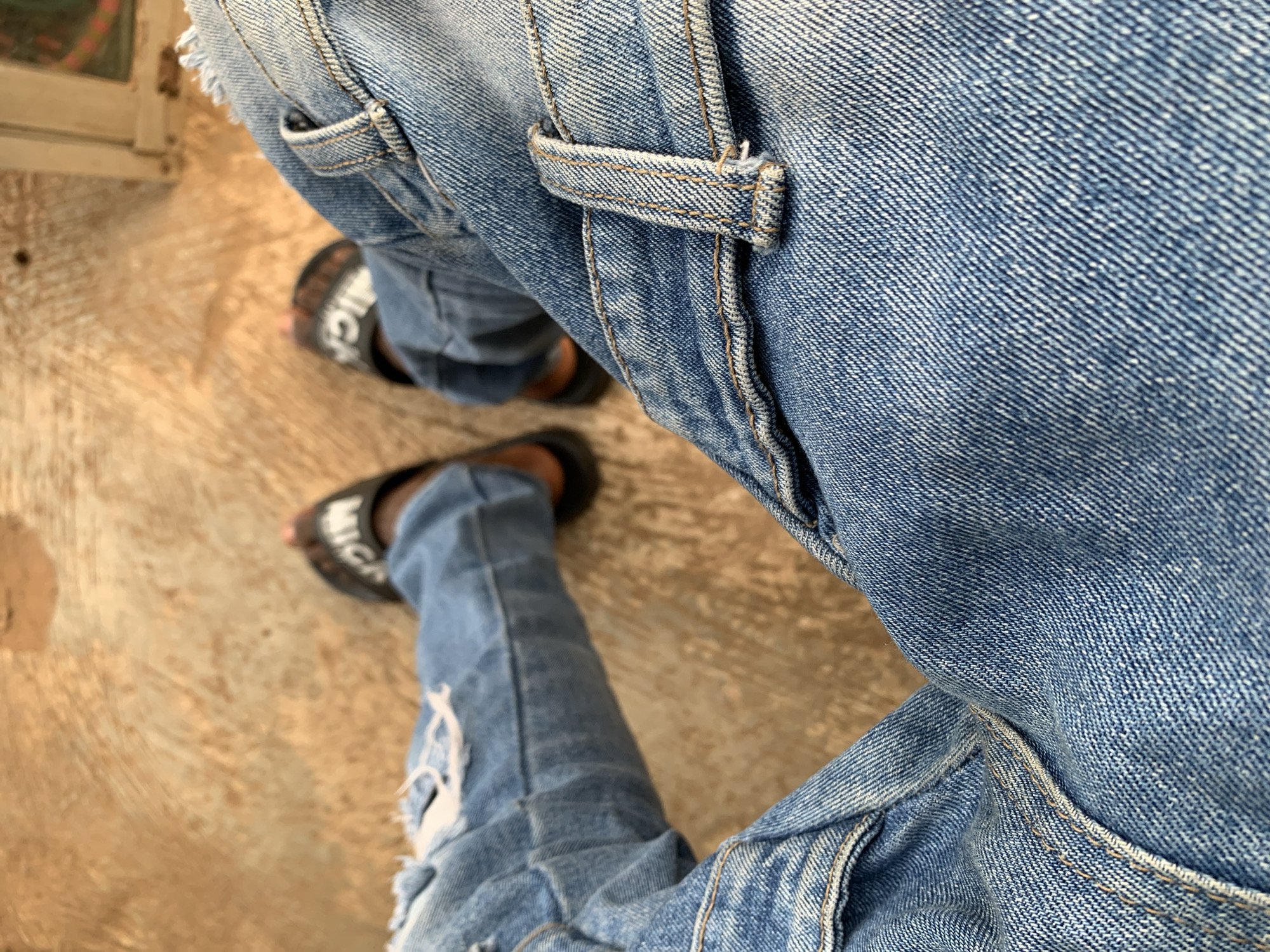 The image presents a high-angle, close-up view of a person's lower body, focusing on their legs and feet. The subject is wearing light blue, distressed denim jeans with visible rips on the left leg, particularly around the knee area. The jeans feature prominent yellowish-brown stitching along seams and a belt loop, highlighting the garment's construction. The denim fabric displays a clear twill weave pattern.
The person is wearing dark-colored slide sandals. On the left sandal, part of a white word, "BALENCI", is visible. On the right sandal, another part of a white word, "MICA", is visible, indicating a brand name, likely "BALENCIAGA".
The background is a light brown or tan ground surface, which appears textured and somewhat worn, possibly concrete or a similar hard outdoor surface. The lighting suggests it is daytime. The overall scene captures a casual moment, with the perspective looking down at the feet. The location context is Kabba, Nigeria.