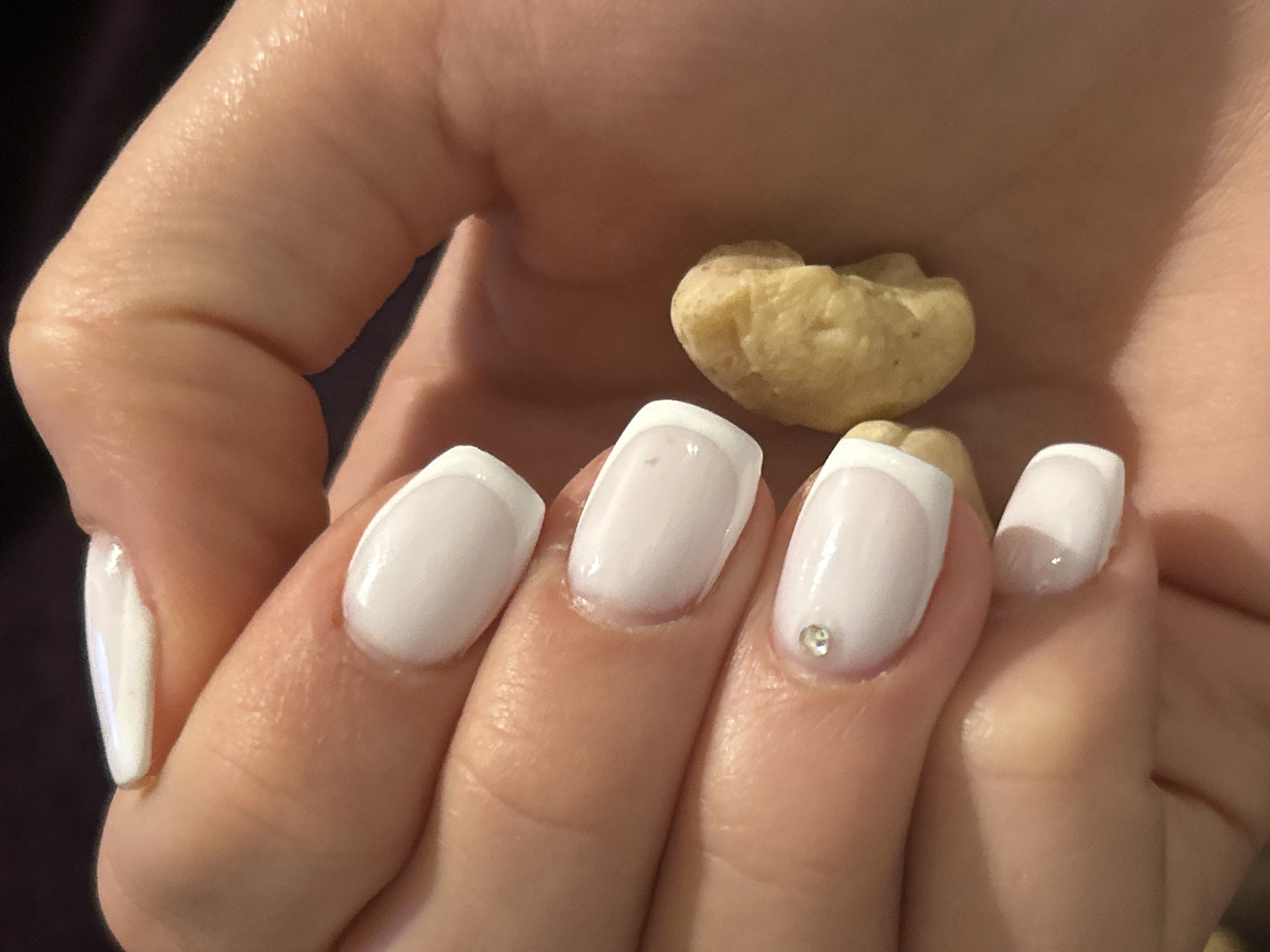 A fair-skinned hand is holding a cashew. The nails are painted in a French manicure style with white tips, and there is a small clear gem on the nail of the ring finger. The background is dark and out of focus.