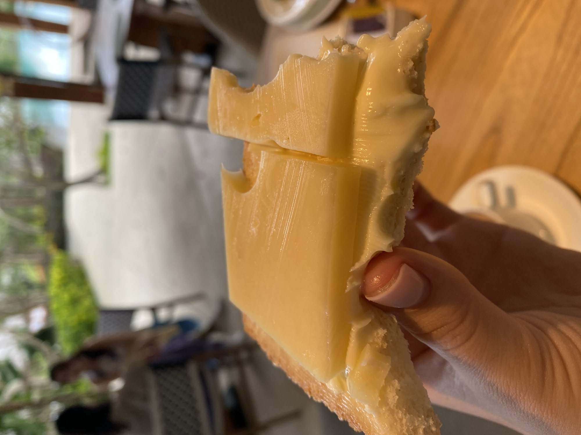 A close-up shot focuses on a slice of bread topped with butter and cheese, held by a person's hand. The bread appears toasted and is spread with a generous layer of melted butter, over which are two slices of yellow cheese. The cheese has a slightly irregular, layered texture. In the background, out of focus, a resort-like setting is visible with greenery, seating areas, and what appears to be a body of water, suggesting the location is Kuta Selatan, Indonesia. Two individuals are partially visible in the background, one seated and another standing, both blurred. The overall lighting suggests an outdoor or well-lit indoor environment.