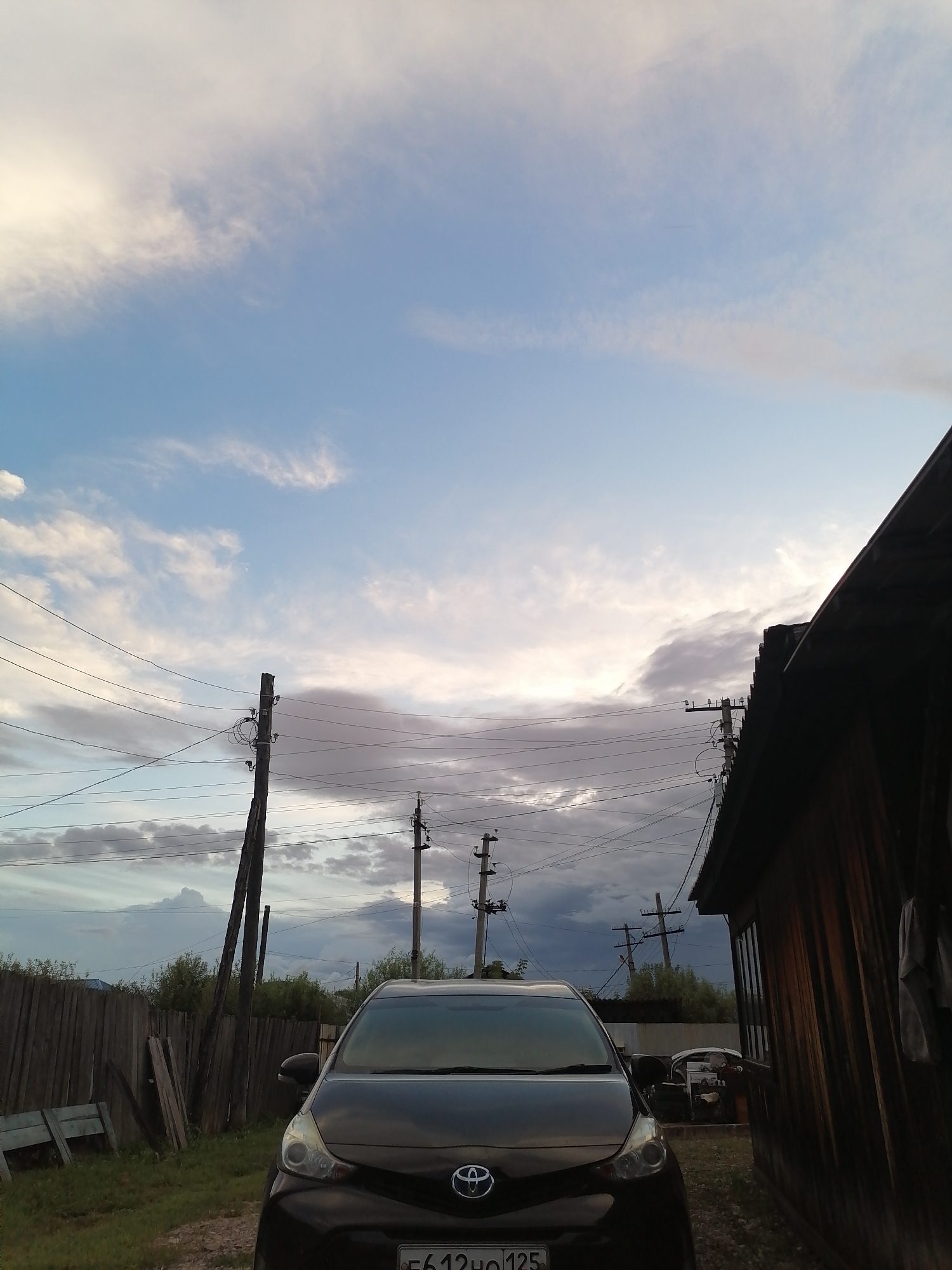 The image depicts a rural scene in Novopokrovka, Russia, under a cloudy sky with patches of blue. In the foreground, a black Toyota hatchback is parked on a dirt and gravel surface. The car's front end, including headlights, grille with a Toyota emblem, and license plate, is clearly visible. To the left of the car is a weathered wooden fence and some discarded wooden planks and a blue bench. In the background, utility poles with numerous wires are prominent, stretching across the sky. To the right, the side of a wooden building with vertical siding is visible, partially obscuring the view. In the mid-ground, more utility poles and vegetation are present. The overall impression is of a residential area in a rural or semi-rural setting.
