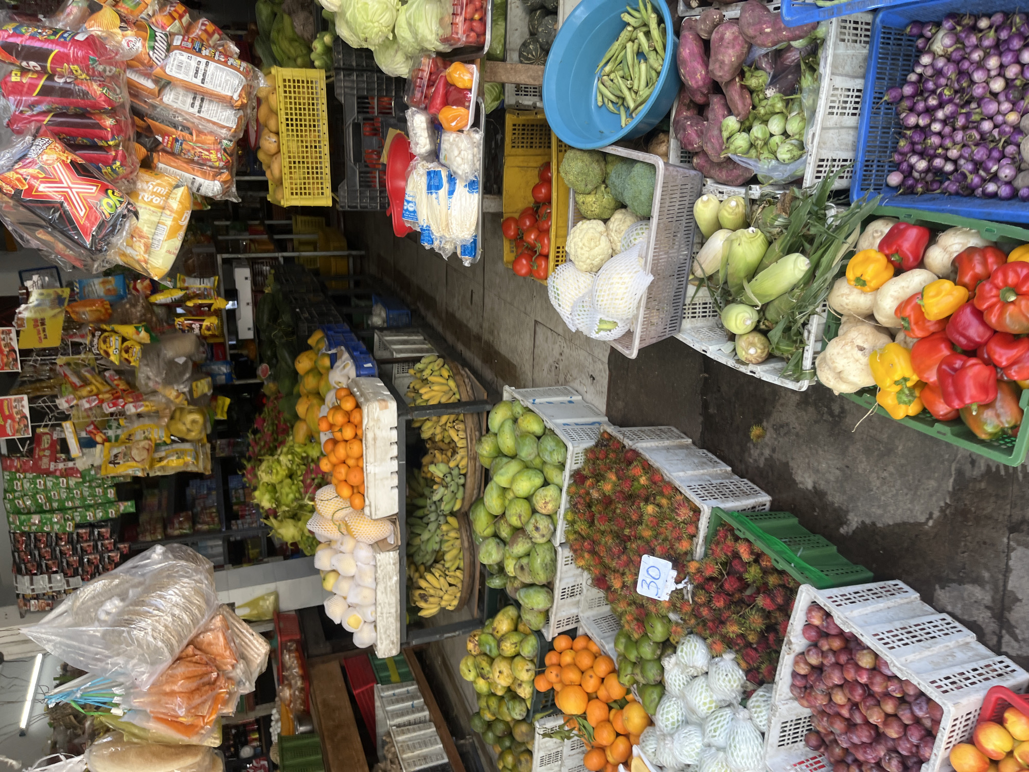 The image depicts an outdoor market stall in the city of Bac Nha Trang Ward, Vietnam. The stall is filled with a variety of fresh produce and packaged goods. In the foreground, crates overflow with fruits and vegetables such as plums, rambutans, oranges, guavas, and what appear to be bell peppers and corn. Behind these, a display of various vegetables including cabbages, tomatoes, sweet potatoes, eggplants, and okra are visible in crates and a blue plastic basin. Packaged snacks and dried goods hang from the ceiling, adding to the bustling market atmosphere. The ground is wet, suggesting recent rain or washing. No people are visible in the image.