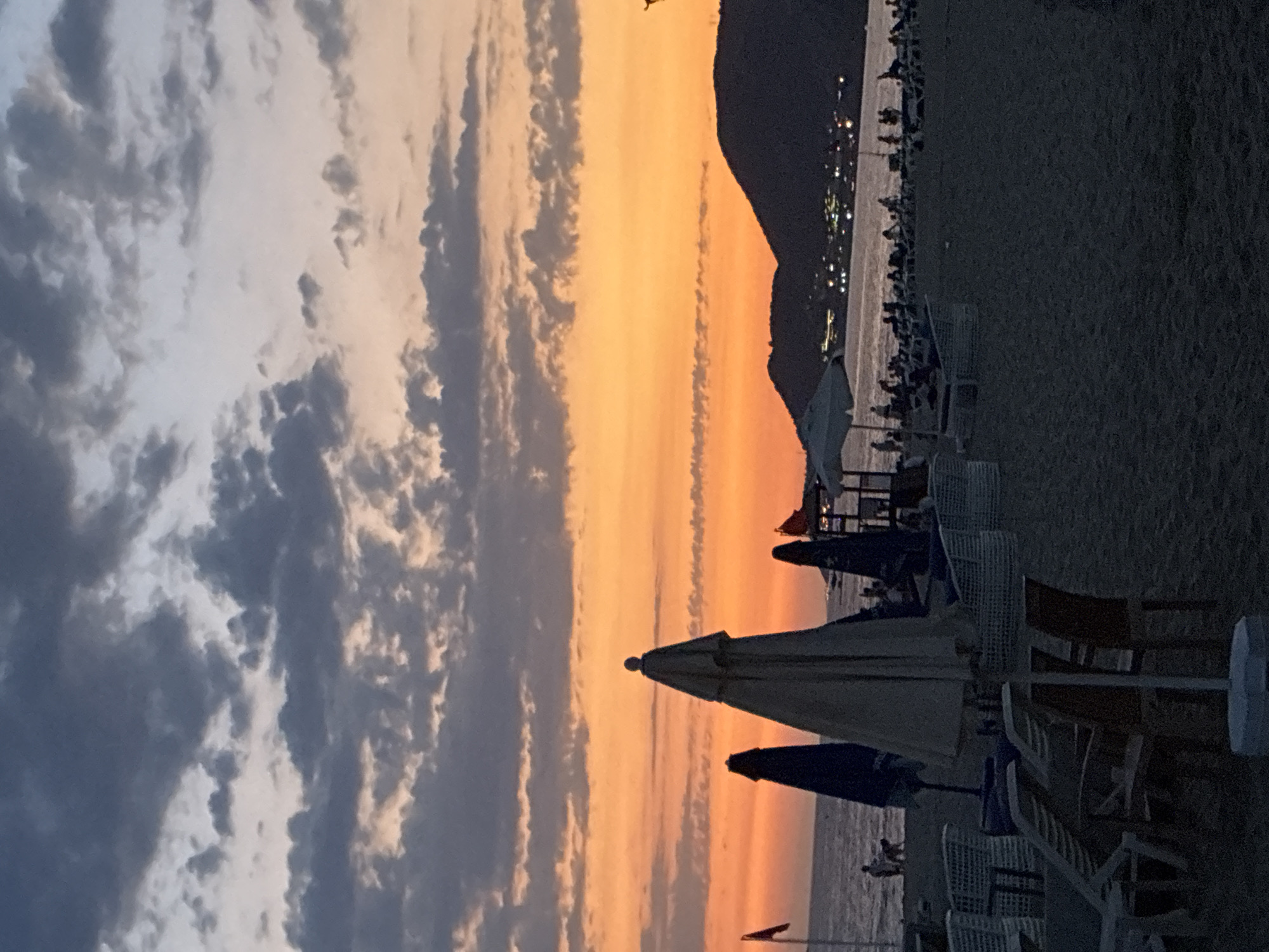 This image depicts a beach scene in Alanya, Turkey, during sunset. The sky is a gradient of orange, yellow, and dusky blue, with scattered clouds. The foreground is dominated by the sandy beach with several beach umbrellas and lounge chairs, some of which are occupied or appear to be arranged for use. In the mid-ground, the sea is visible, with a dark silhouette of a coastline and hills in the background, dotted with lights indicating a town or city. Several figures, including some walking and others gathered, are present on the beach and near the water. The overall atmosphere is that of a peaceful evening at a seaside resort.