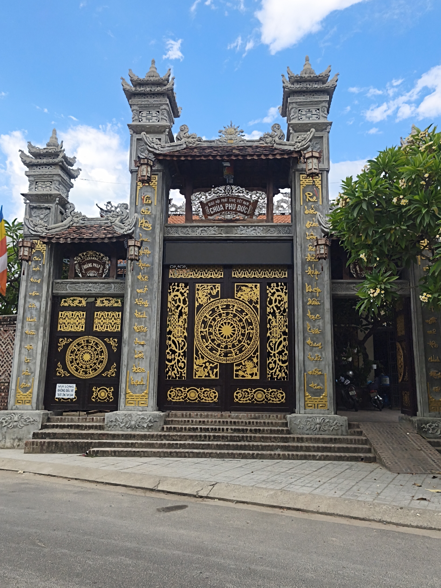 A wide shot captures the ornate entrance to Phu Duc Pagoda in Nha Trang, Vietnam. The pagoda gate is grand, featuring large, dark wooden doors adorned with intricate golden circular motifs, possibly representing mandalas or Buddhist symbols. Flanking the doors are tall, grey stone pillars decorated with golden calligraphy. These pillars are topped with pagoda-style roofs and decorative carvings, matching smaller decorative structures on either side of the main entrance.
Above the central gate, a sign reads "GIAO HOI PHAT GIAO VIET NAM CHUA PHU DUC," identifying it as a temple of the Vietnam Buddhist Sangha, Phu Duc Pagoda. Smaller signs and decorations are visible on the left, including one that says "VUI LÒNG KHÔNG ĐẬU XE TRƯỚC CỔNG TAM QUAN" (Please do not park vehicles in front of the main gate). A vibrant Vietnamese flag is partially visible on the far left.
A lush green tree with small white flowers grows on the right side of the entrance, partially obscuring the view into the pagoda grounds. Through an opening in the gate on the right, motorcycles and what appear to be people can be seen, suggesting the pagoda is active. The ground leading up to the entrance is paved with stone and brick, with several stone steps. A concrete ramp runs alongside the steps for accessibility. The sky above is bright blue with scattered white clouds, indicating it is daytime. The overall impression is one of a well-maintained, culturally significant religious site.