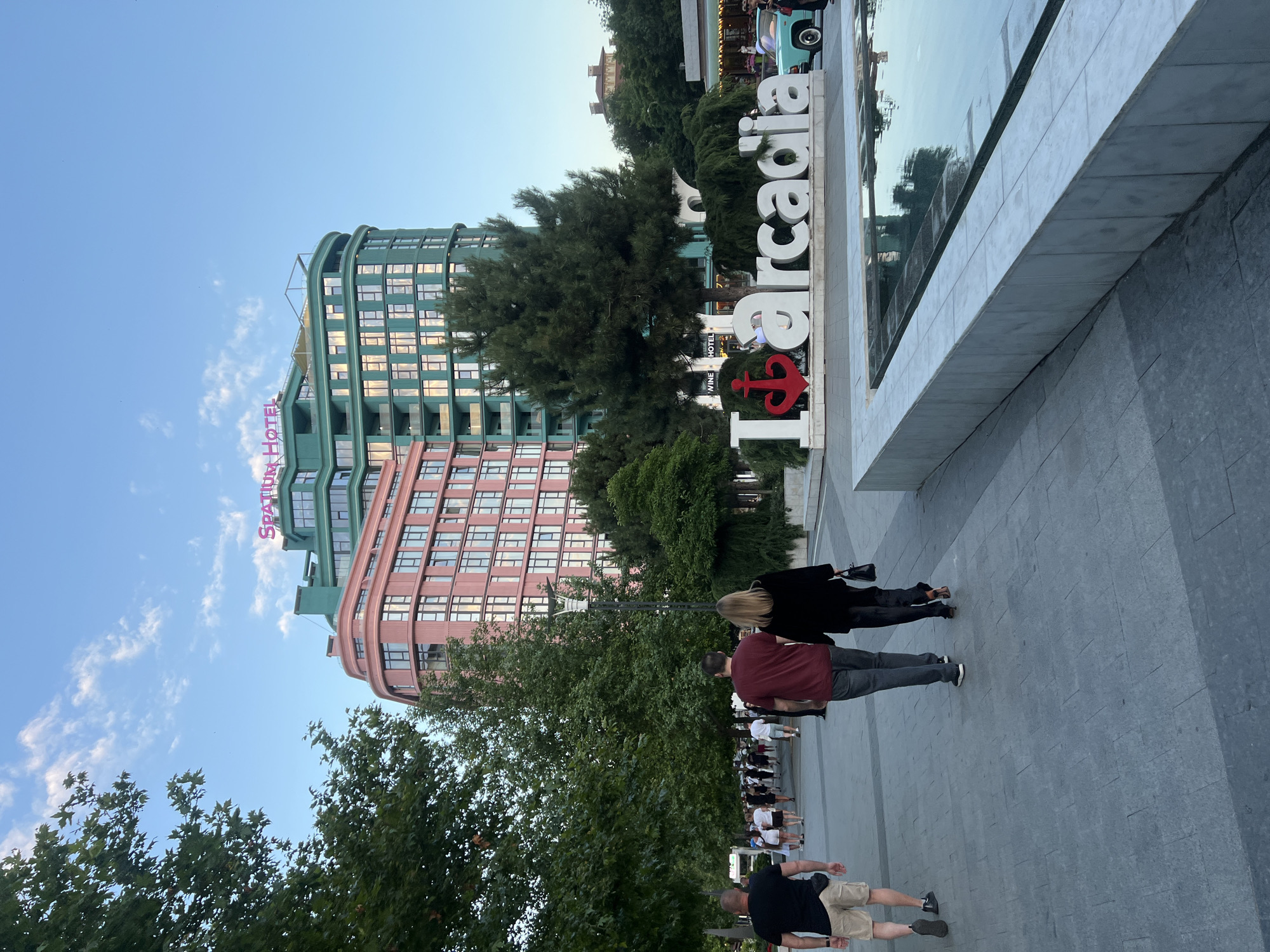 The image depicts the Spatium Hotel in Arcadia, Odesa, Ukraine, on a clear day with a blue sky and scattered clouds. In the foreground, a wide paved walkway is visible, with several people walking towards the viewer's left. Two individuals, a man in a burgundy t-shirt and a woman in a black jacket, are in the center of the frame, walking away from the camera. To the left, a man in a black t-shirt and beige shorts is also walking away. A large, three-dimensional sign spelling "I Arcadia" with a red anchor replacing the letter "A" is prominently displayed in the middle ground. Behind the sign, lush green trees and a light blue vintage car can be seen. The Spatium Hotel itself is a striking structure with two distinct colored sections: one a soft pink and the other a vibrant green, both featuring multiple floors with many windows and balconies. The hotel's name, "SPATIUM HOTEL," is illuminated in pink on its rooftop. The overall scene conveys a lively atmosphere in an urban recreational area.