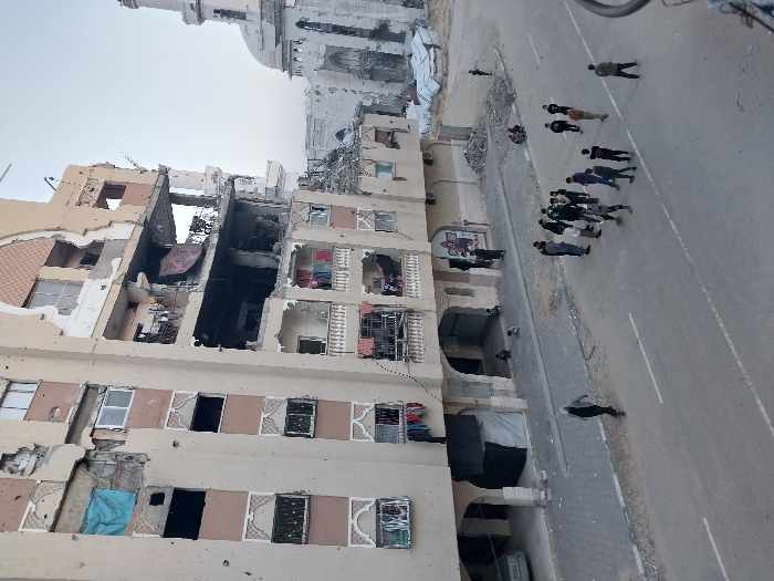 This image depicts a damaged urban environment in Khan Yunis, Palestinian Territory. The primary subject is a multi-story, light-colored building on the left, exhibiting extensive structural damage including large missing sections of exterior walls, exposed interior rooms, shattered windows, and numerous pockmarks consistent with impacts. A blue tarp covers a window opening on a lower floor, and a large red piece of fabric hangs from a damaged balcony on an upper floor. Laundry is visible hanging on several balconies.
Approximately 20-25 individuals are present. A group of about 15-20 men and youths are gathered in the center of the asphalt street, which features faded white lane markings. One individual walks alone on the left side of the street. Additional individuals, including children, are seated on the sidewalk near the damaged building. A silver car is partially visible parked on the left.
In the background, to the right, a large white mosque with a prominent dome and a minaret is visible, also showing significant structural damage and debris. Piles of rubble are present on the sidewalks and at the base of the buildings. The sky is clear and light-toned.