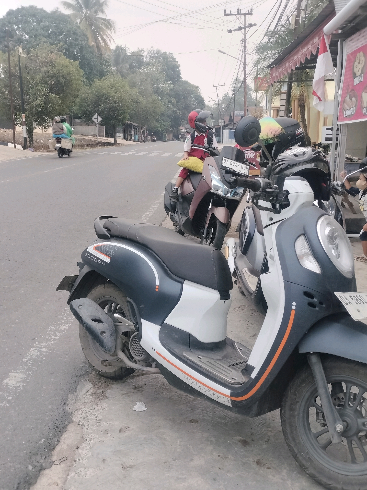 A wide shot captures a street scene in Pembataan, Indonesia, where several motorbikes are parked alongside a road. In the foreground, a black and white scooter with orange accents is prominently displayed. Behind it, another scooter, a darker color with a rider in a red shirt and helmet, is parked. Further down the road, a third scooter, green in color with two passengers, is seen moving away from the camera. The environment appears to be a bustling urban area, with trees lining the street and buildings visible in the background. The sky is overcast, suggesting a cloudy day.