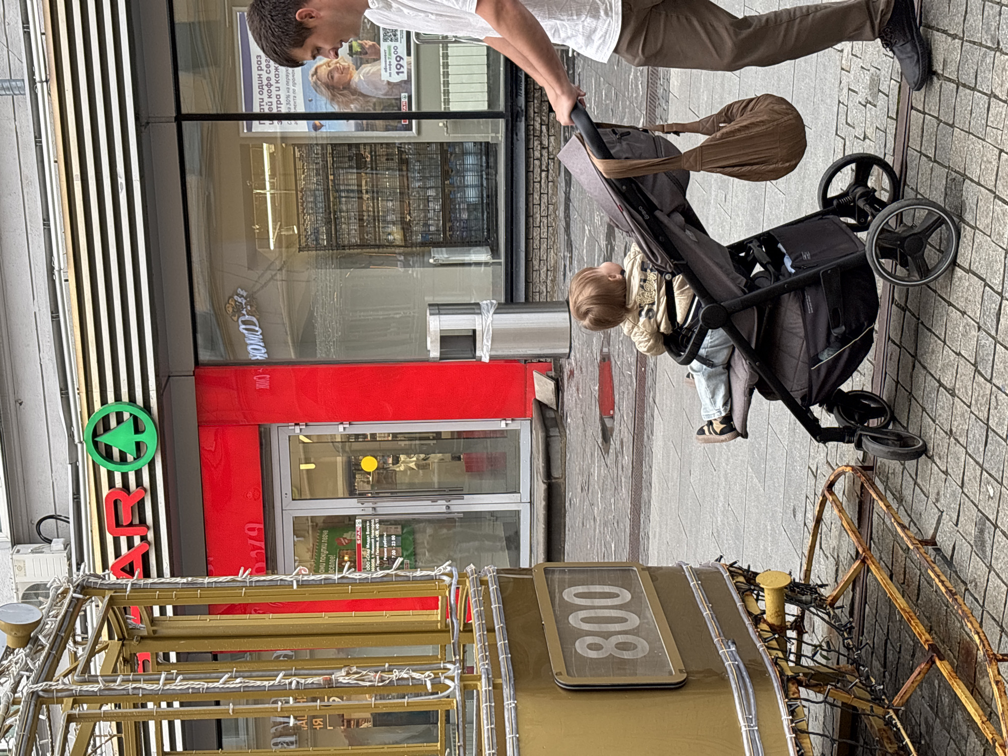 In Nizhny Novgorod, Russia, a man is pushing a baby in a stroller on a cobblestone street next to a vintage tram labeled "800". The tram is a golden color and appears to be decorated with lights. In the background, there is a red building with a SPAR grocery store logo and store entrance. A poster featuring a woman holding a coffee cup is displayed in the store window. The weather appears to be overcast, and the street shows signs of recent rain with puddles on the ground. The man is wearing brown pants and a white shirt, and the baby is dressed in a beige jacket.